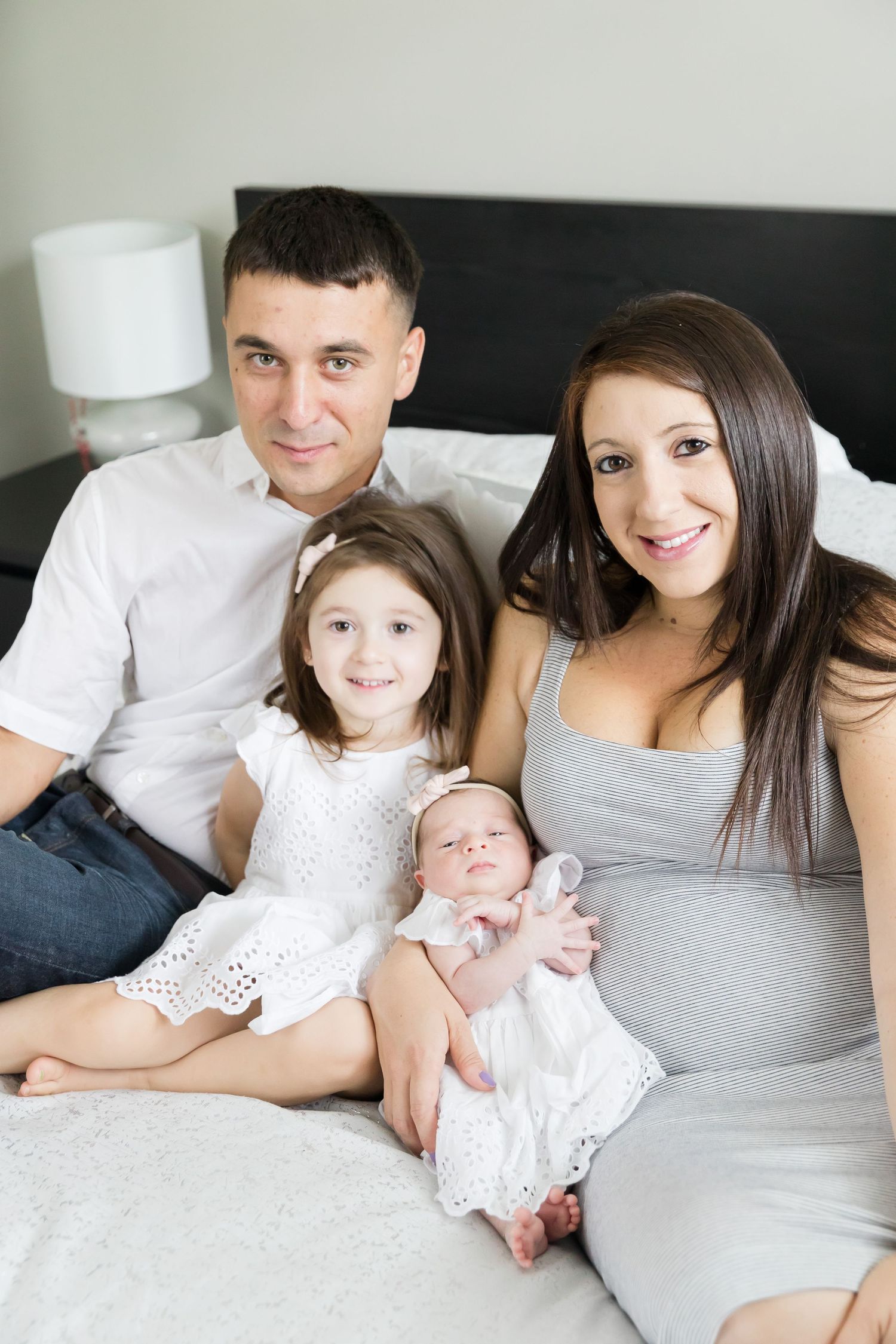Family posing together on bed with young children wearing white dresses.