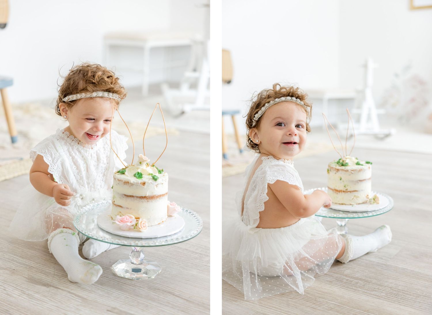 Toddler in white lace dress and headband sits by a small decorative birthday cake on cake stand.