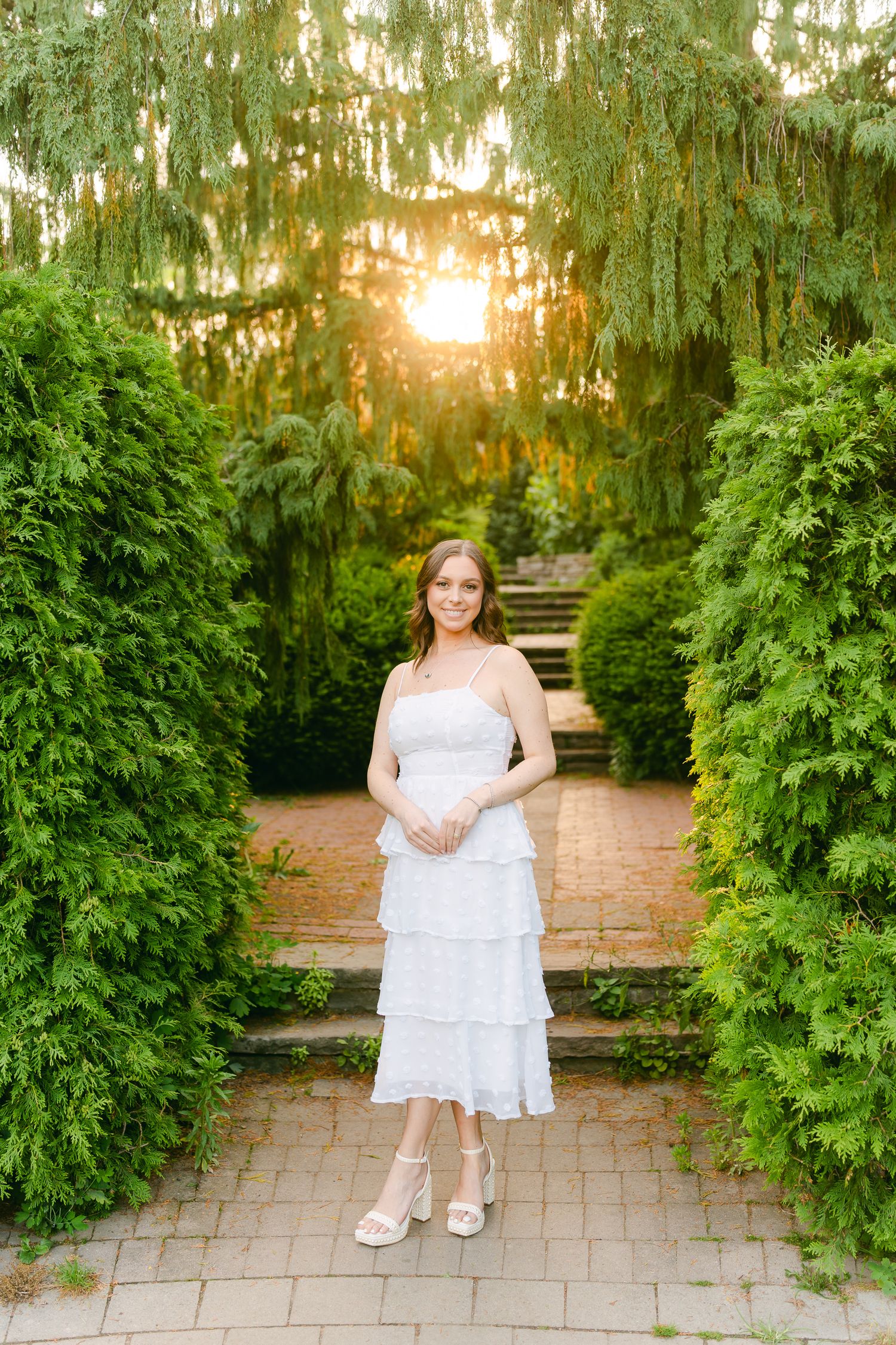 Woman in white tiered dress poses on garden steps with golden sunlight filtering through trees.