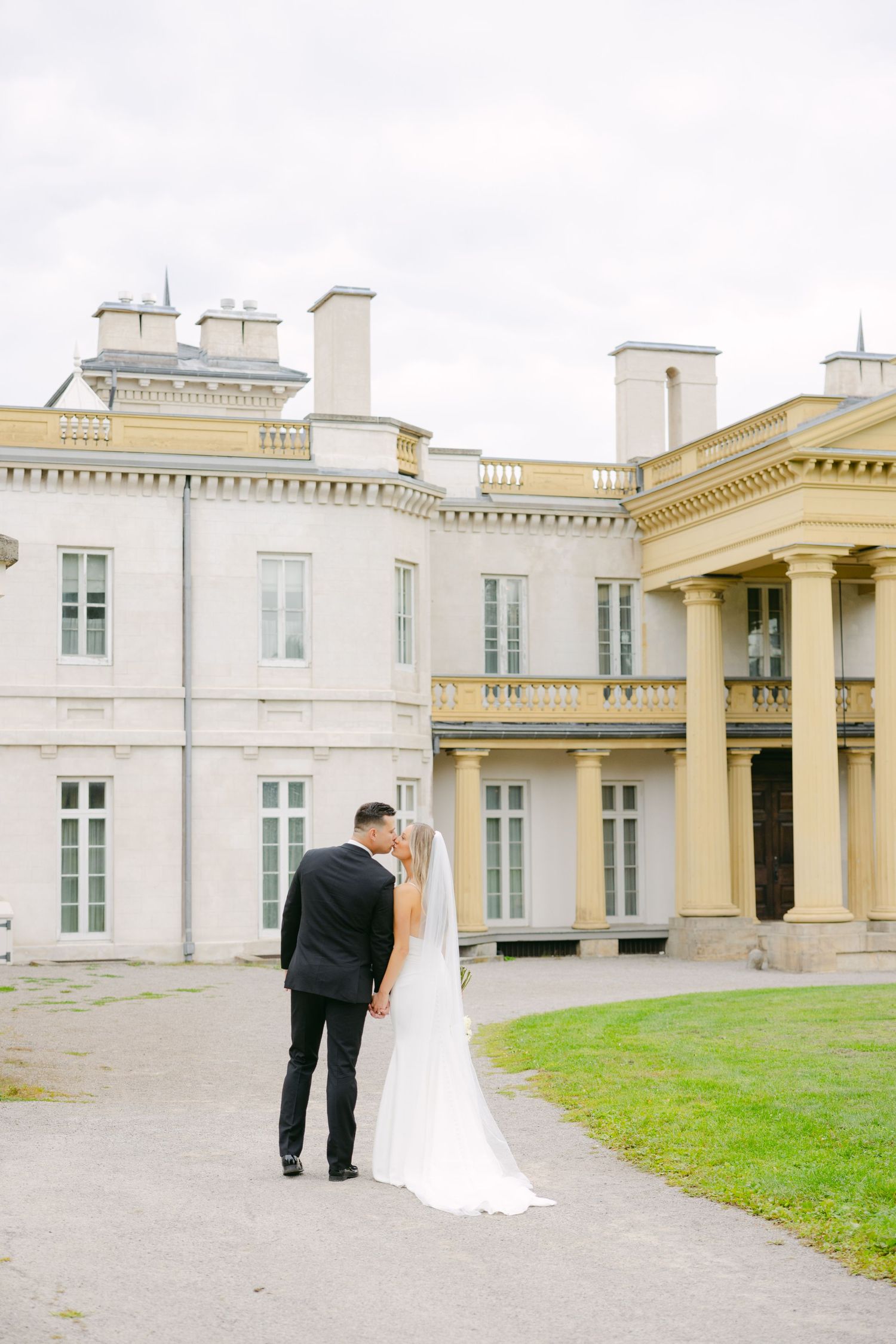 Wedding couple stands in front of elegant white mansion with neoclassical architecture and manicured lawn.