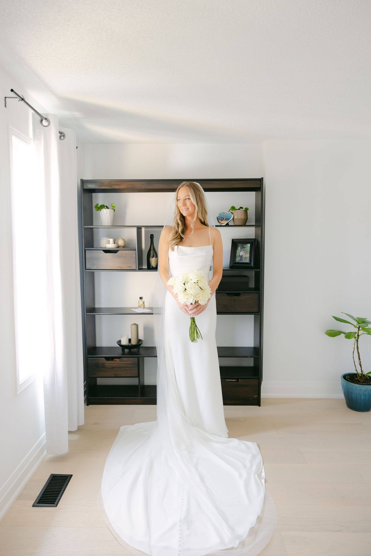 A classic wedding dress with long train is showcased in a bright room with built-in shelving.