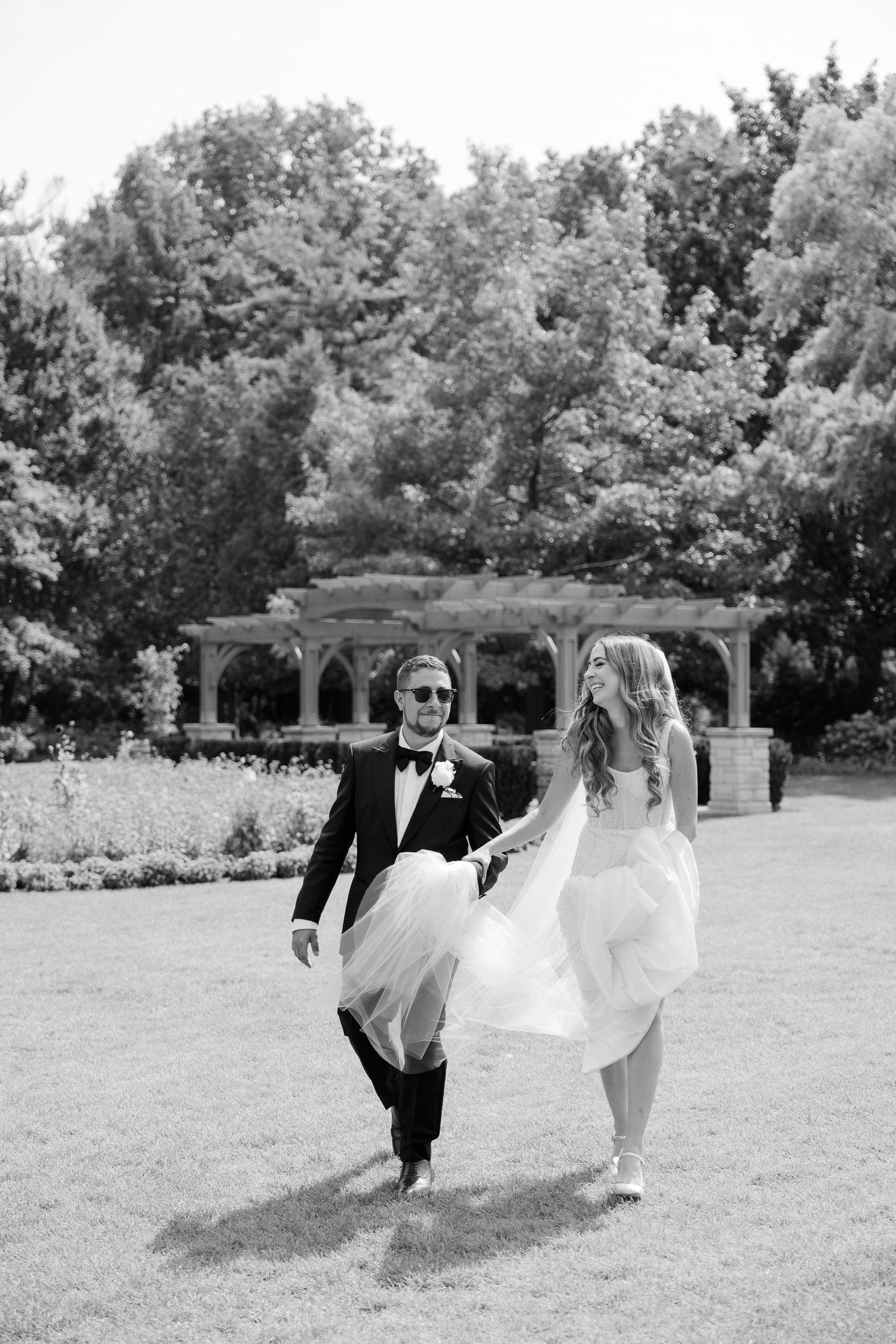 A joyful couple in wedding attire playfully walks across a manicured lawn with a pergola in the background.