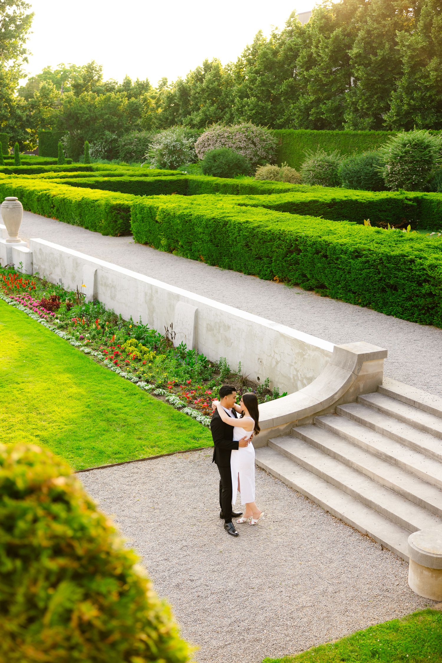 A romantic moment unfolds on garden steps surrounded by vibrant green hedges and blooming flowers.