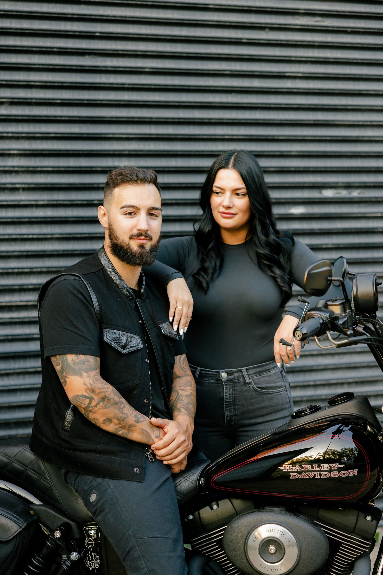 A couple in black attire poses with their motorcycle against a corrugated metal wall.