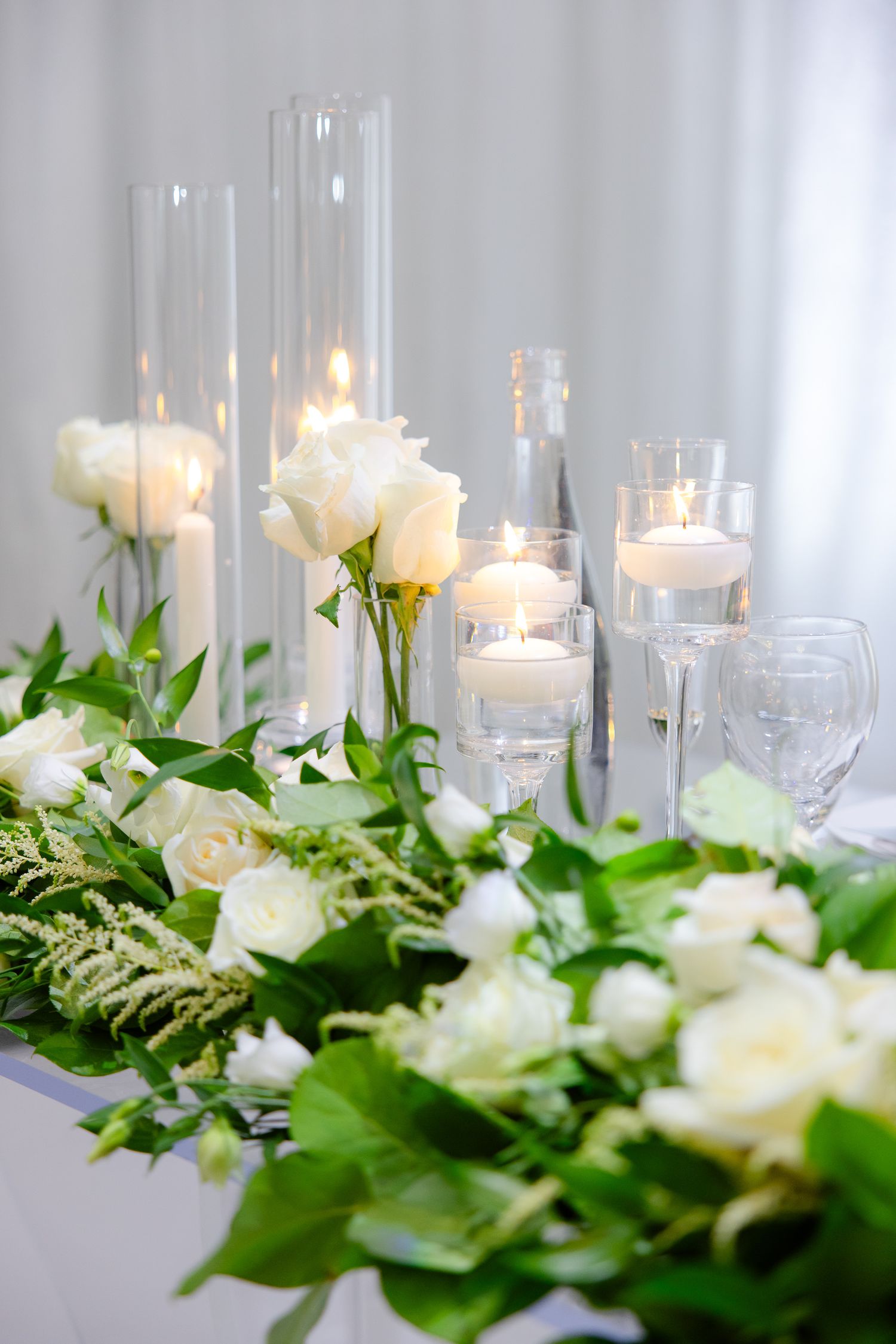 Romantic wedding table decor with white roses, greenery and glass candle holders creating soft lighting.