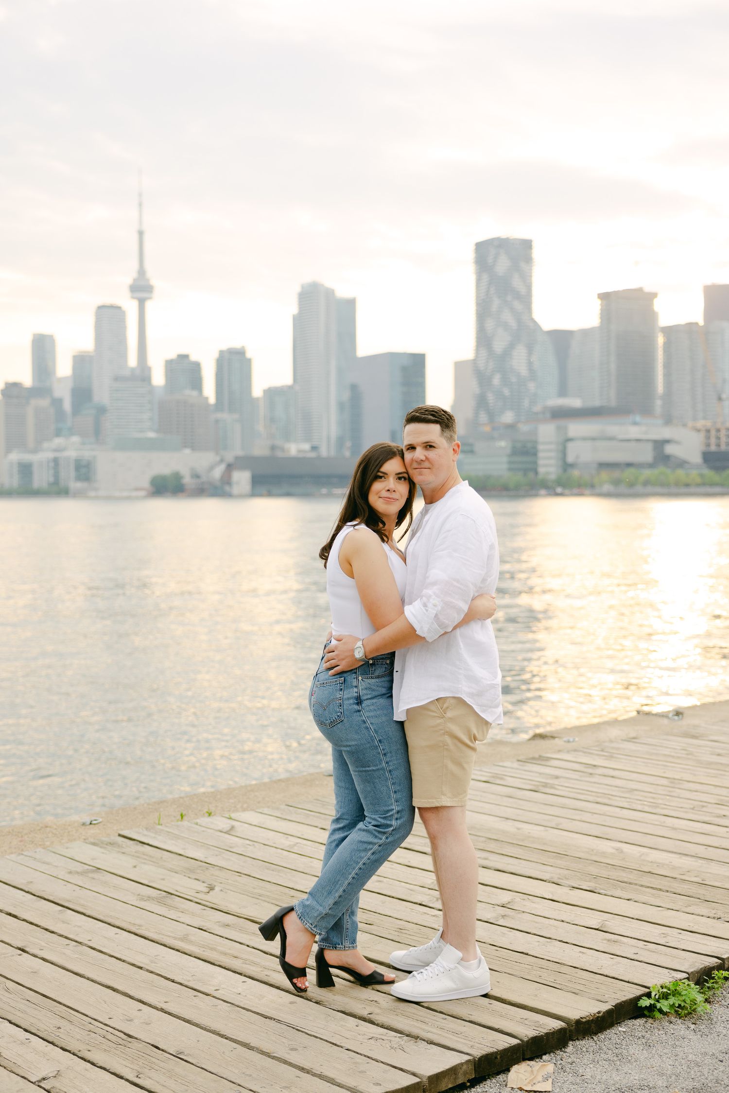 Couple embracing on waterfront boardwalk with Toronto cityscape in soft evening light.