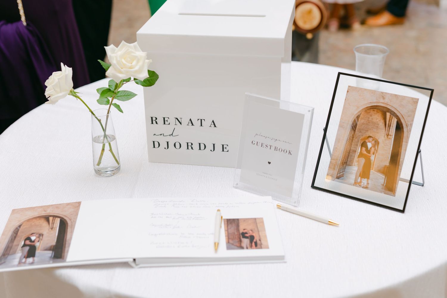 Wedding guest book table with white roses, framed photos, and name cards in an elegant setup.