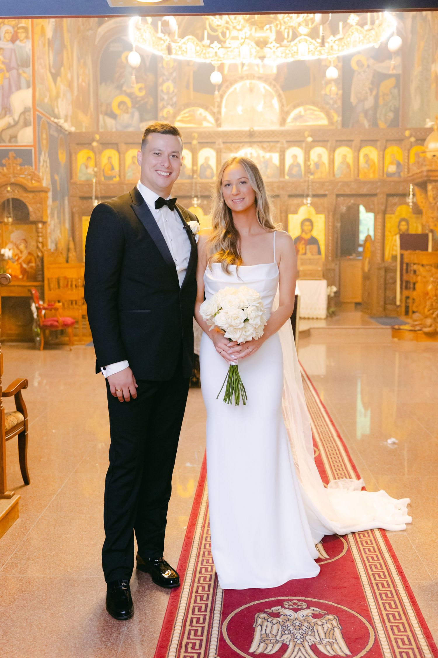 Elegant wedding portrait in Orthodox church with golden iconostasis and chandelier in background.