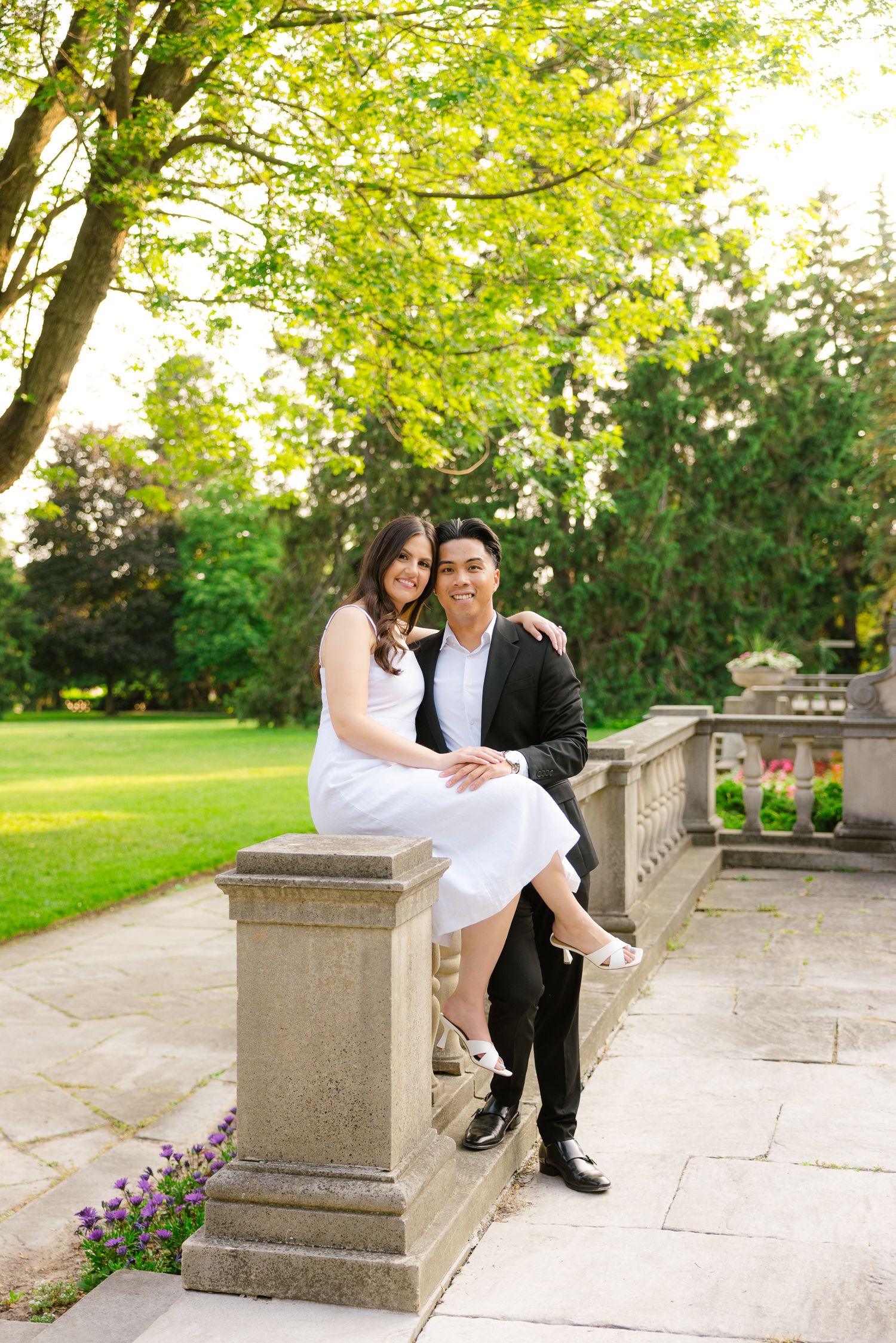 Couple in formal attire sits on stone balustrade in sunlit garden setting.
