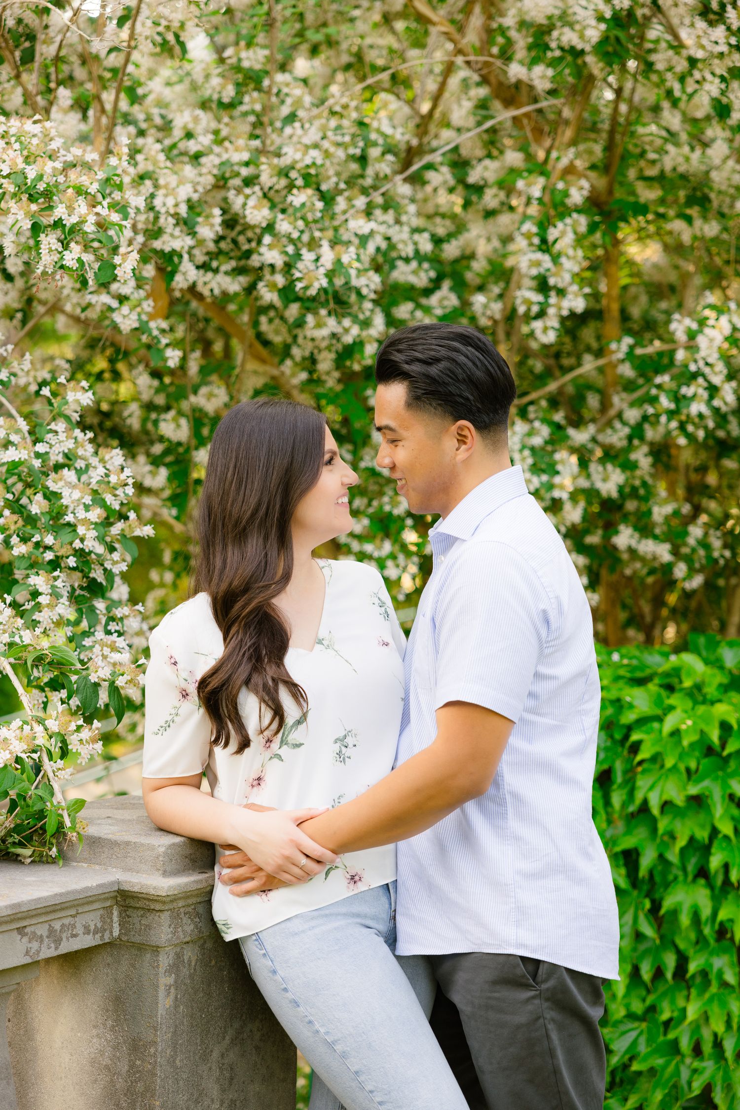 Romantic couple embracing near stone railing with flowering trees in background.