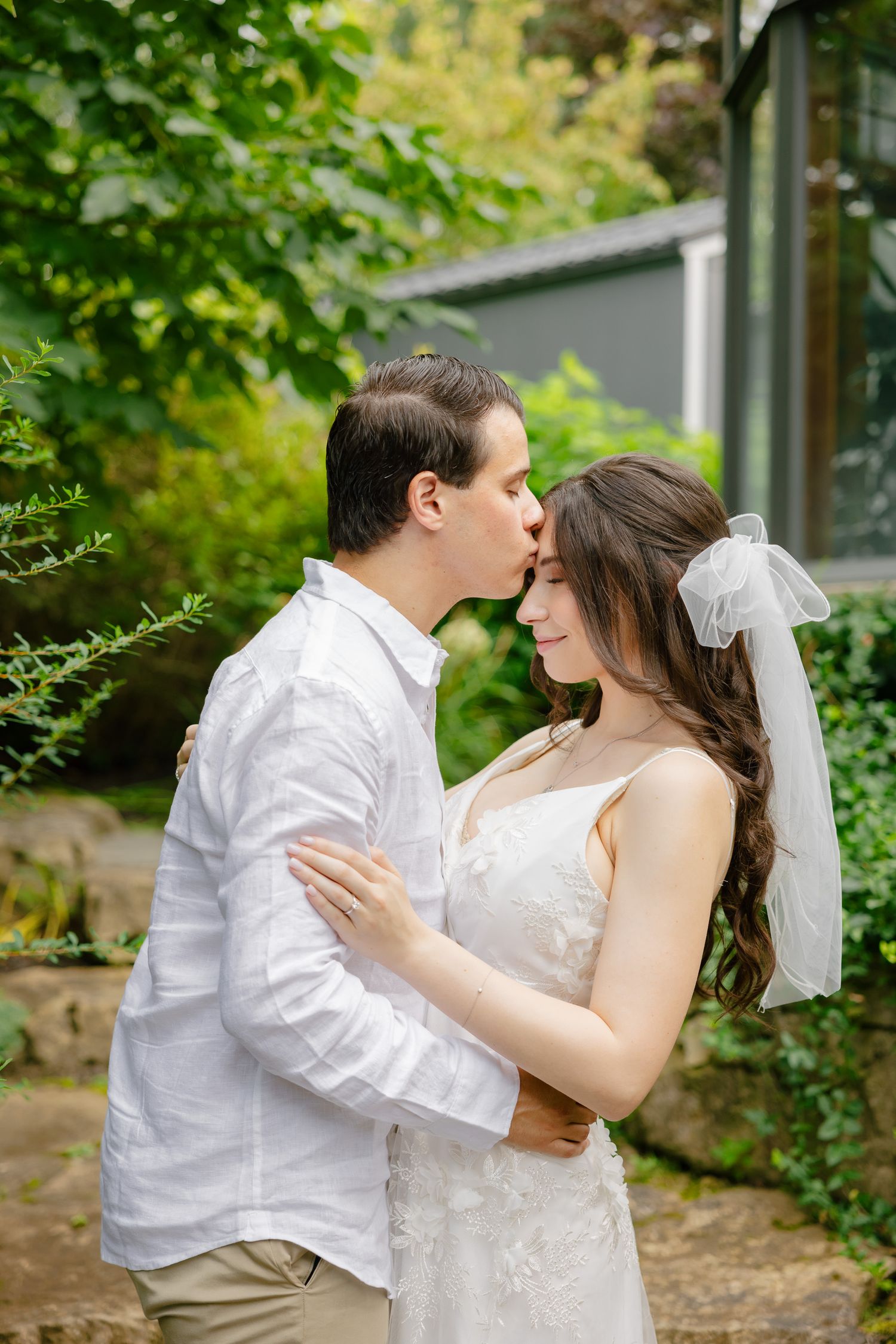 Sweet wedding moment captured in a lush garden setting with greenhouse background.
