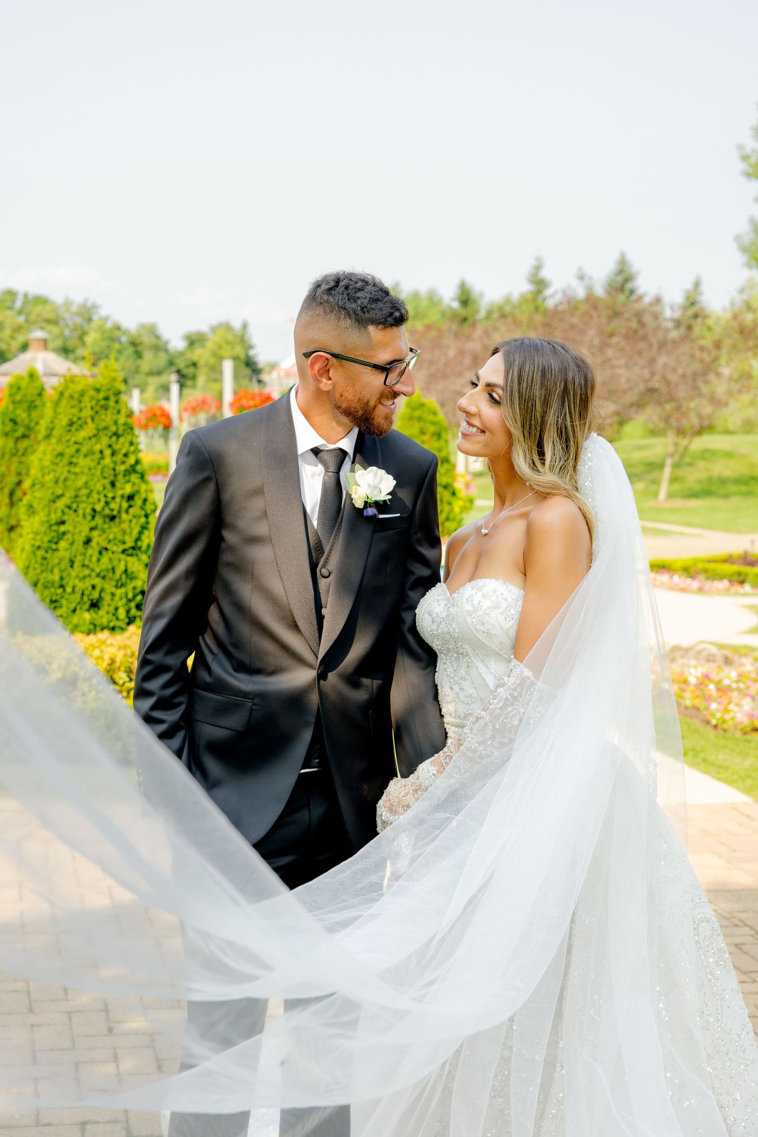 Wedding photo capturing romantic moment as veil flows in breeze among garden evergreens.