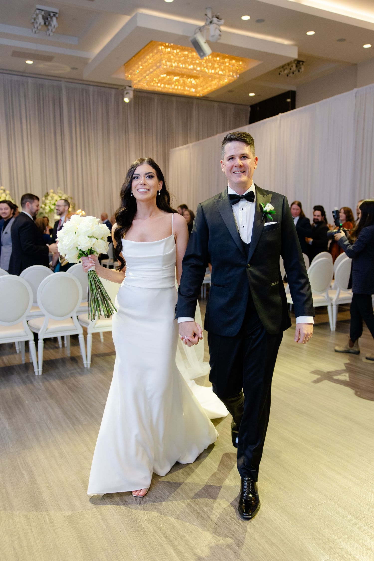 Newlyweds walk down the aisle in an elegant ballroom with crystal chandelier.