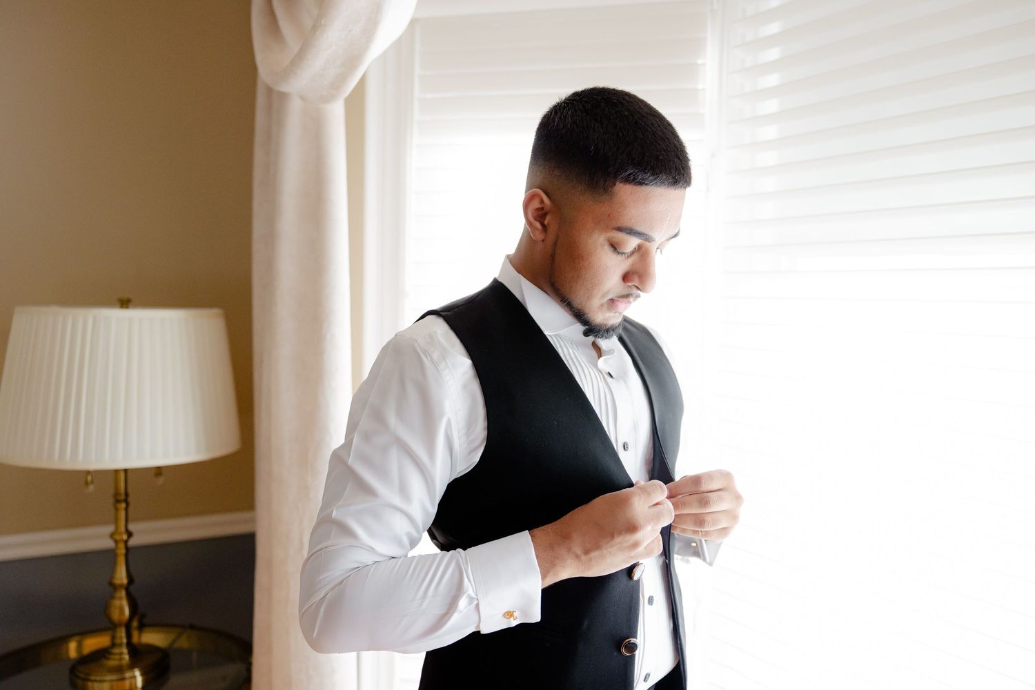 A person fastens buttons on their formal wear while getting ready near a window with soft lighting.