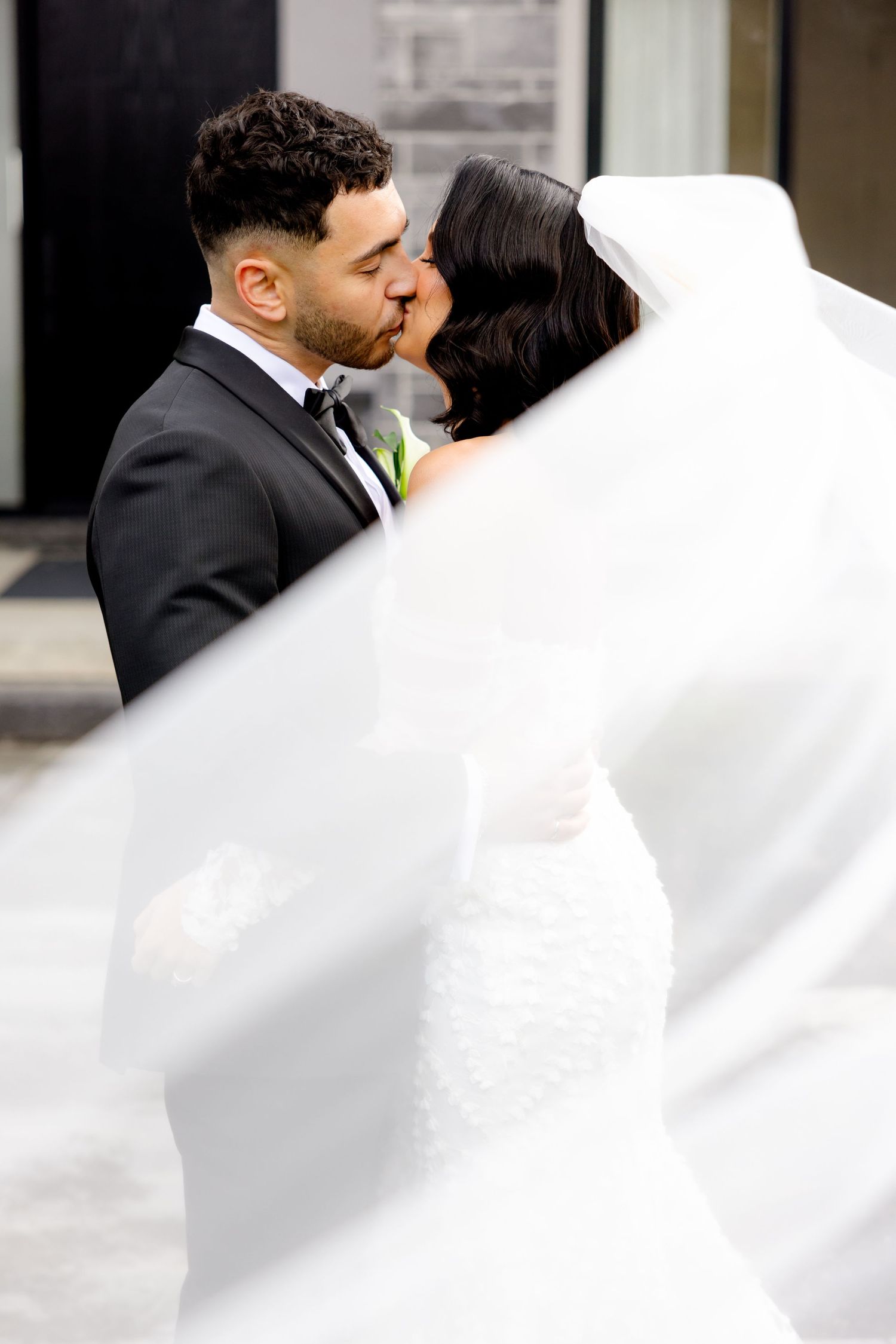 Newlywed couple sharing a romantic kiss as the bride's veil flows in the breeze outside a modern venue.