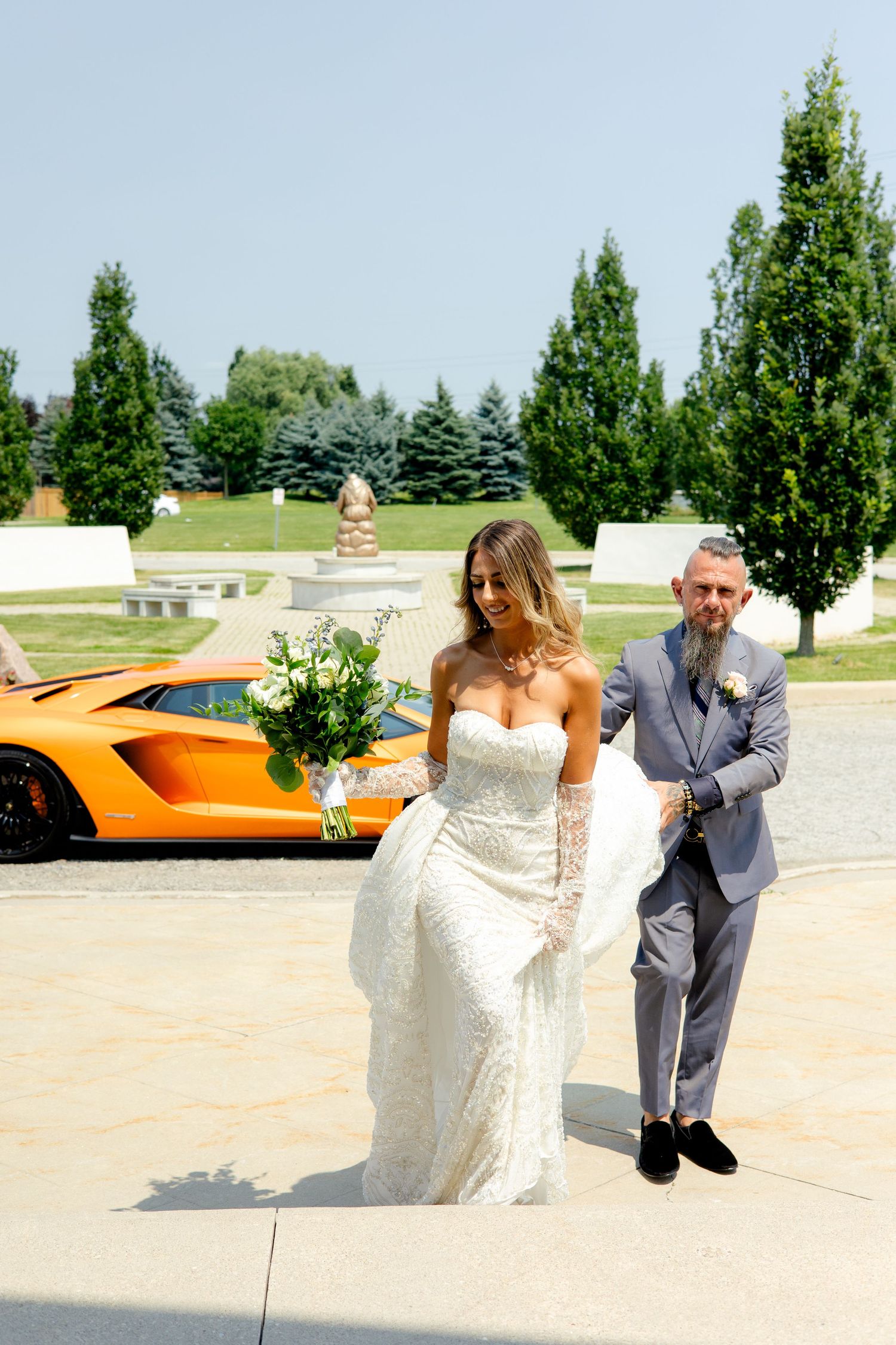 A wedding couple strolls along a paved walkway with tall trees and an orange sports car in the background.