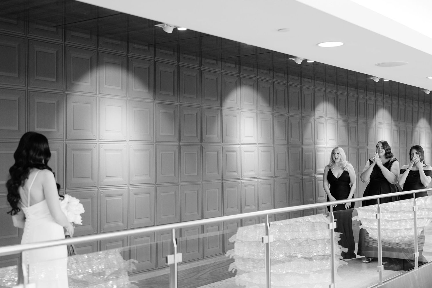 A black and white photo of bridesmaids reacting emotionally to seeing the bride in a modern hallway.
