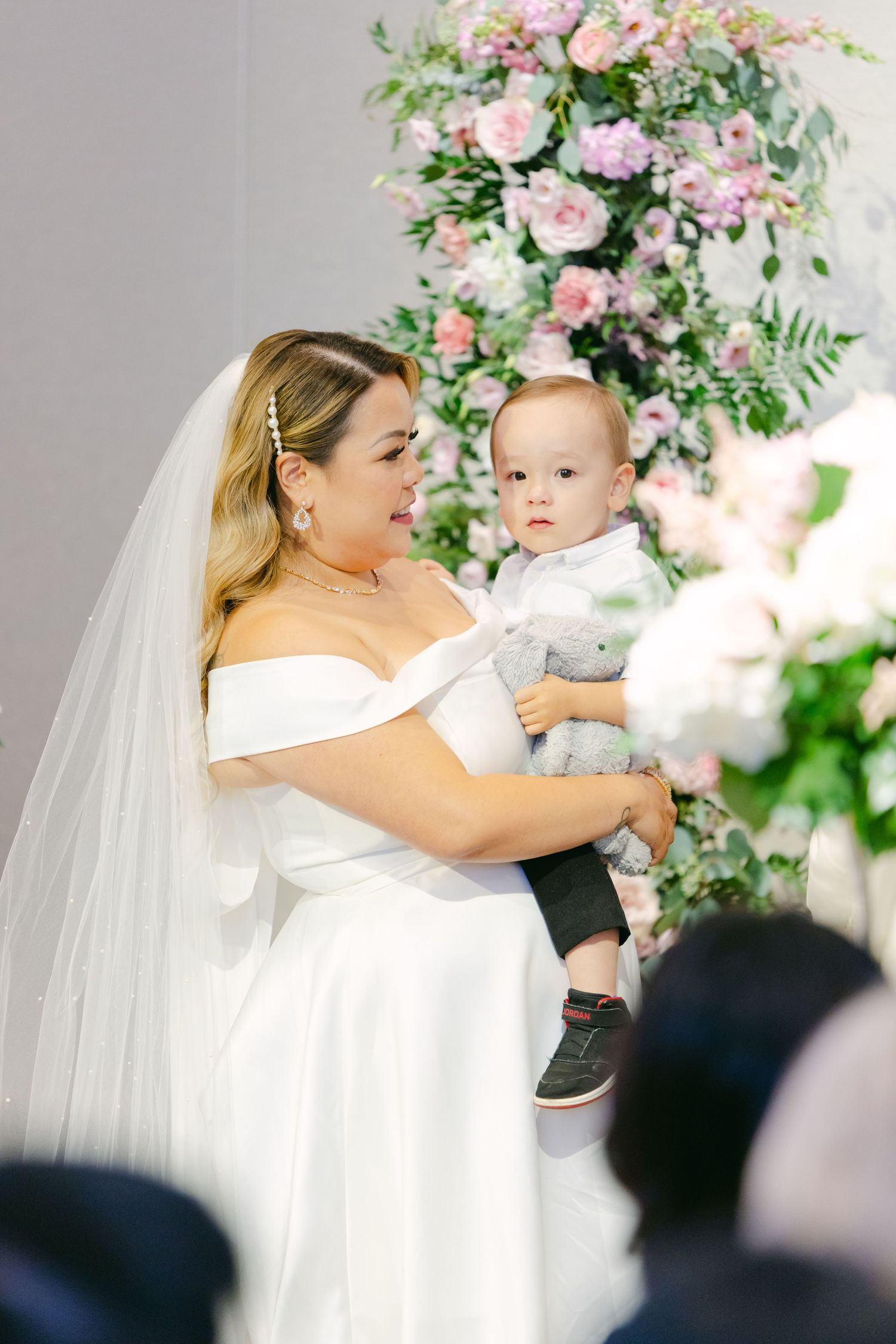 Bride in off-shoulder gown holds small child during wedding ceremony.