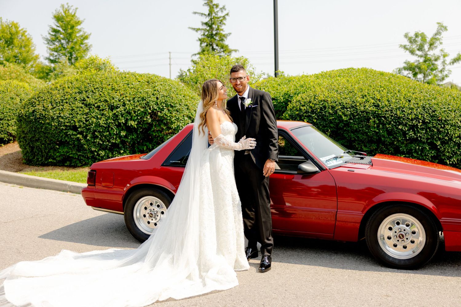 Happy couple celebrates their wedding day next to a red vintage car surrounded by green landscaping.