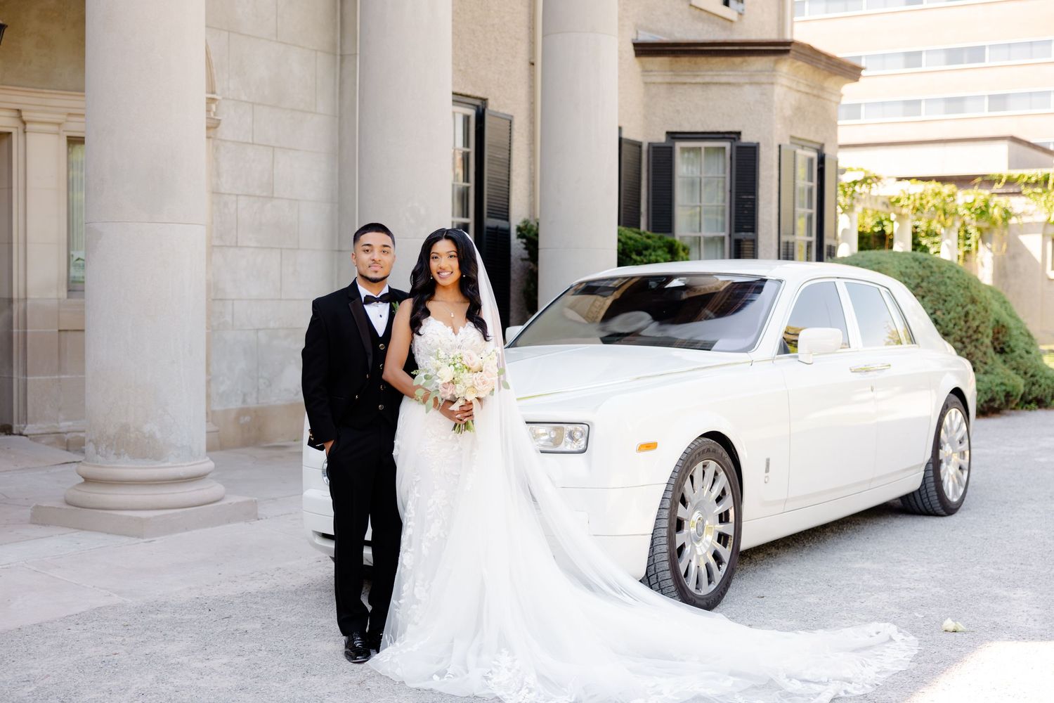 A couple poses beside a luxury white Rolls Royce car in front of a building with elegant columns.