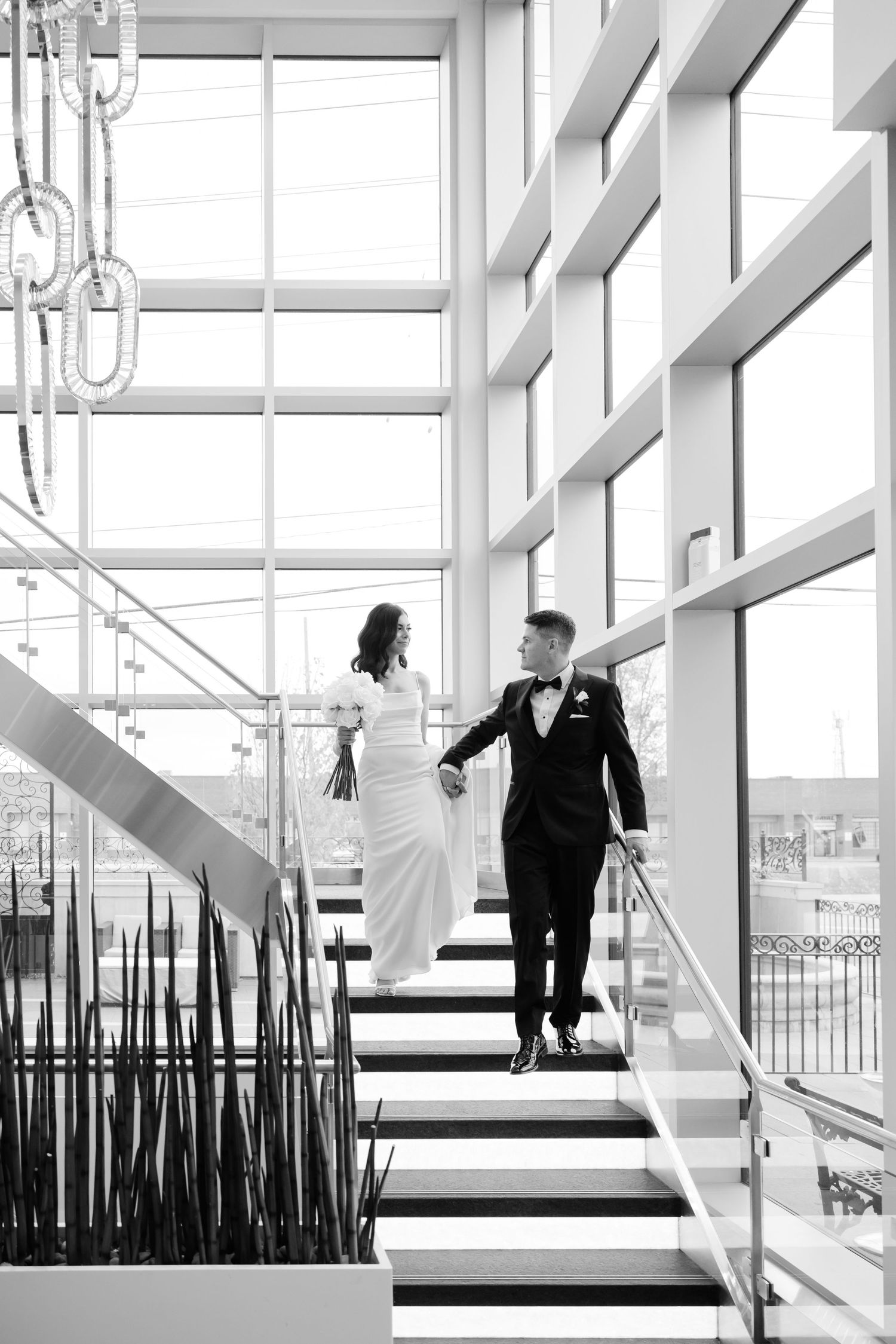 Wedding couple ascending modern white staircase with floor-to-ceiling windows.