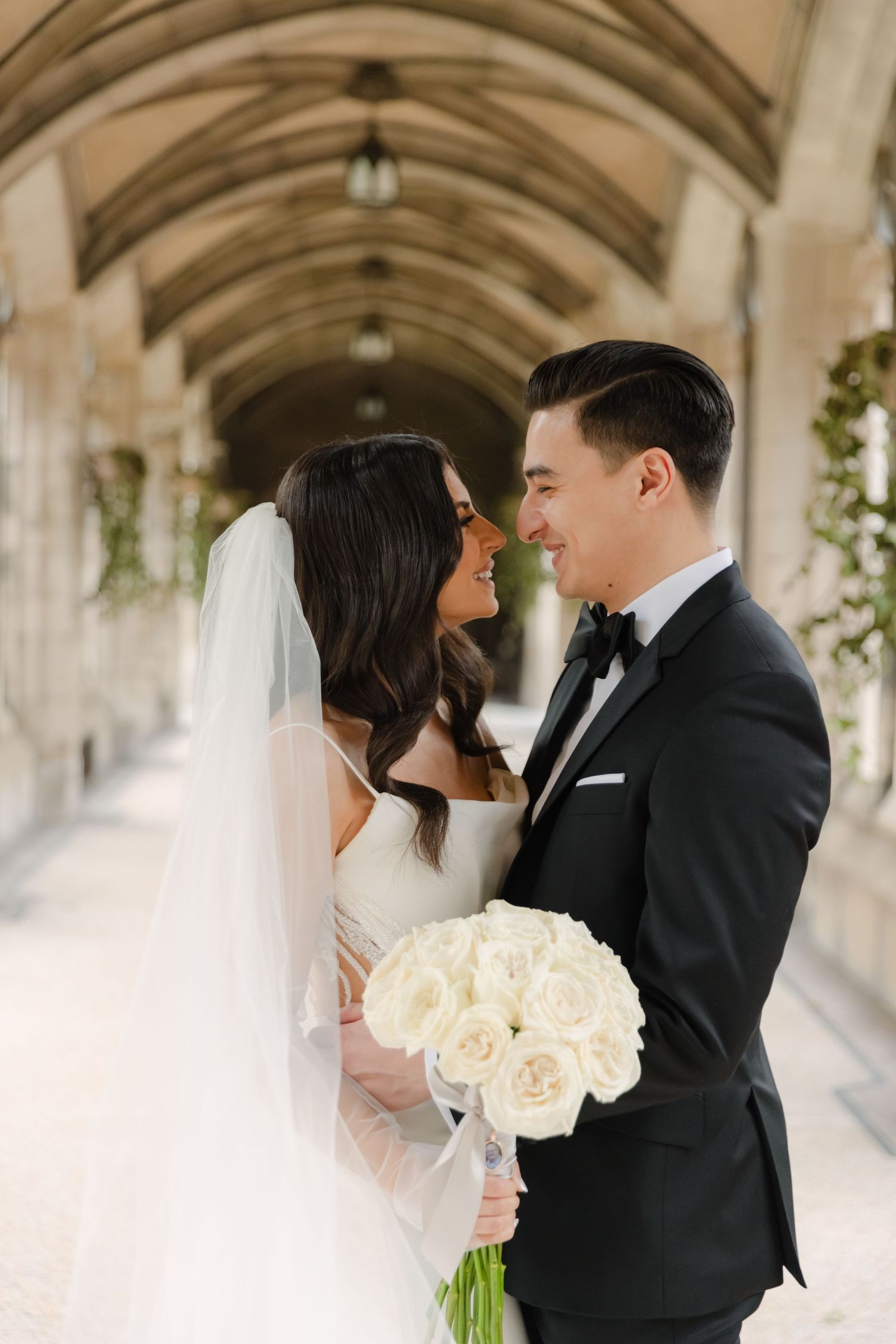 Bride holds white rose bouquet while sharing intimate moment in Gothic style hallway.