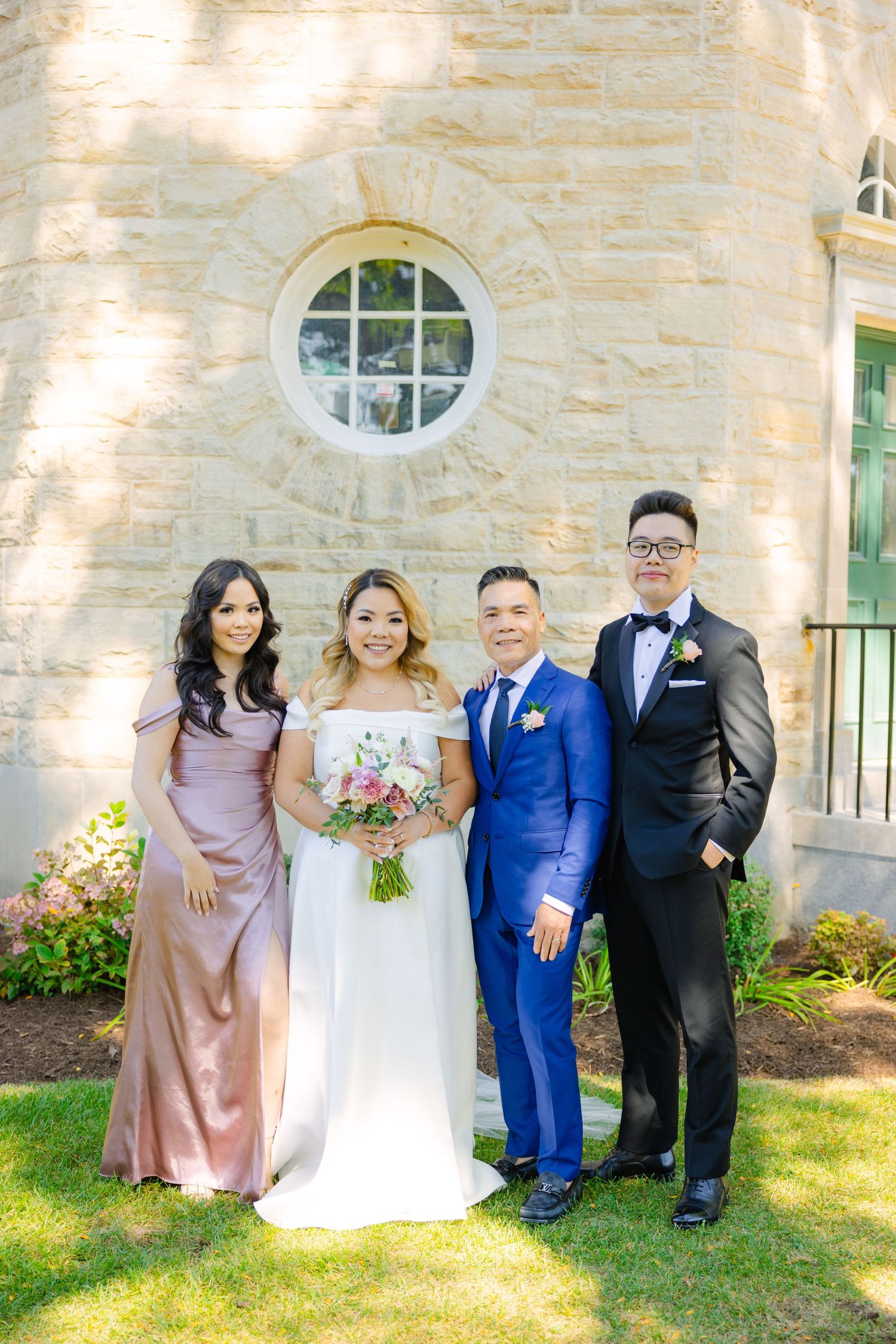 Formal wedding party group stands against stone wall with circular window in elegant attire.