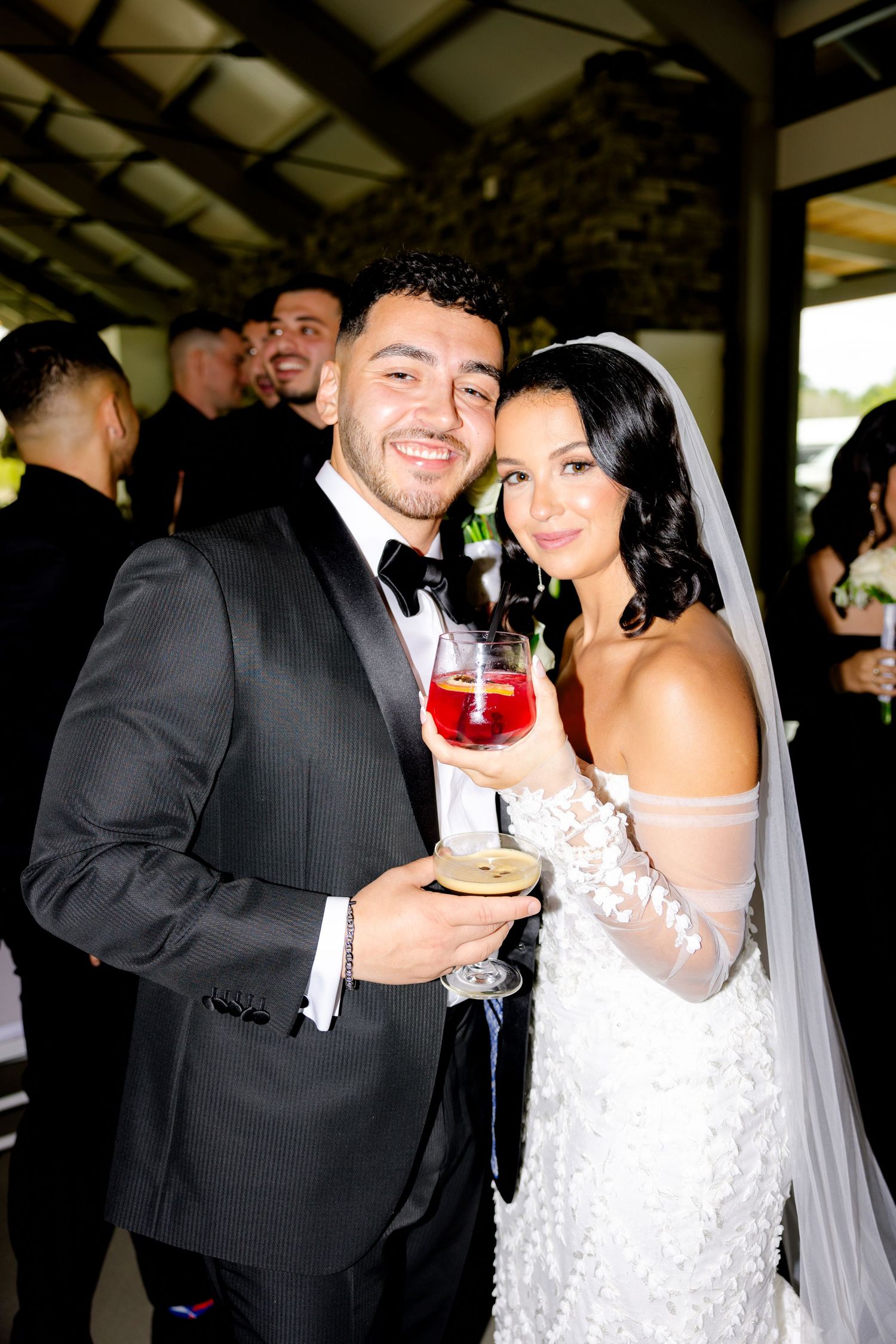 A bride in a white lace gown and groom in a black tuxedo toast with cocktails at their wedding reception.