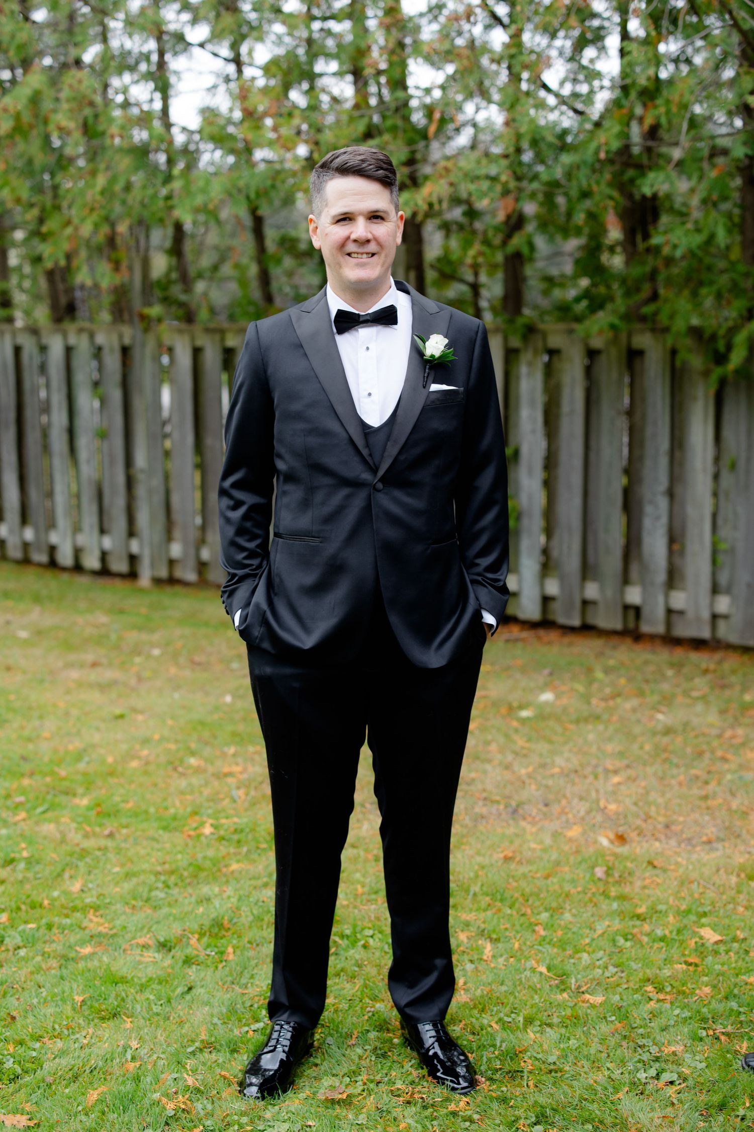 A formal portrait of someone in a classic black tuxedo with white boutonniere against a garden backdrop.