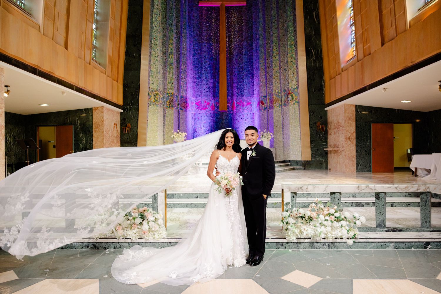Wedding portrait with flowing cathedral veil against purple-lit church backdrop.