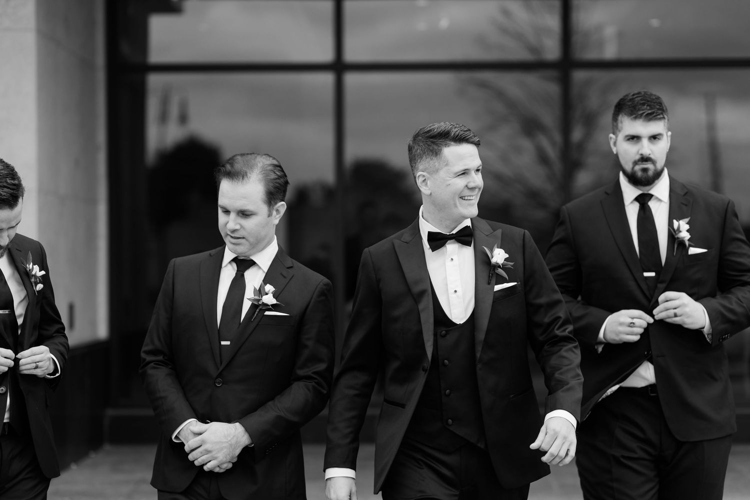 Black and white photo of groomsmen in tuxedos sharing a candid moment outside.