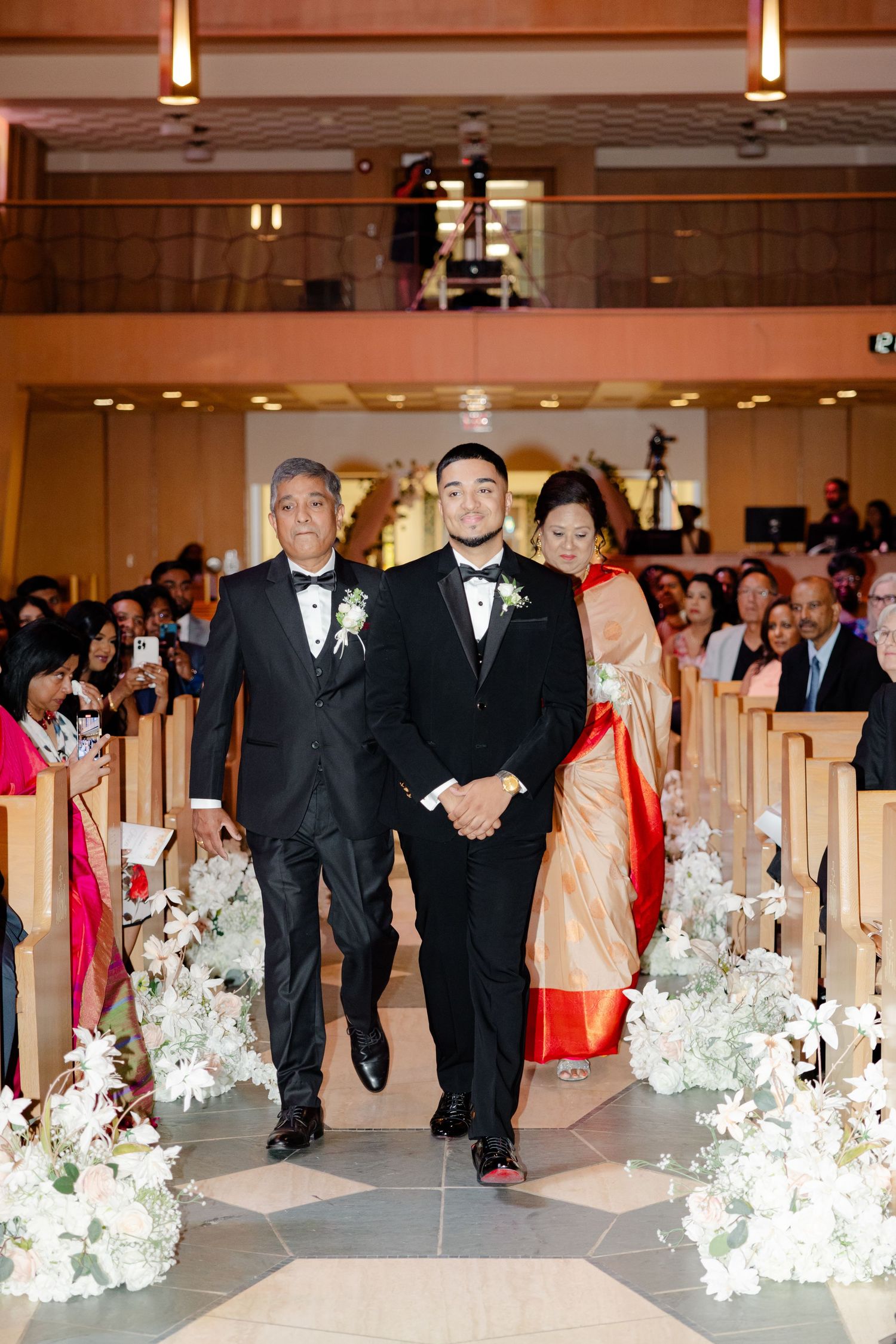 Wedding party members walk down decorated church aisle during ceremony processional.