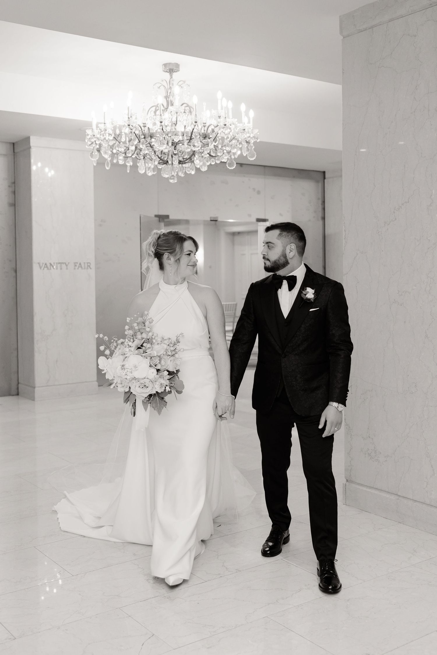 A black and white photo captures a wedding couple walking through an elegant venue with chandeliers.