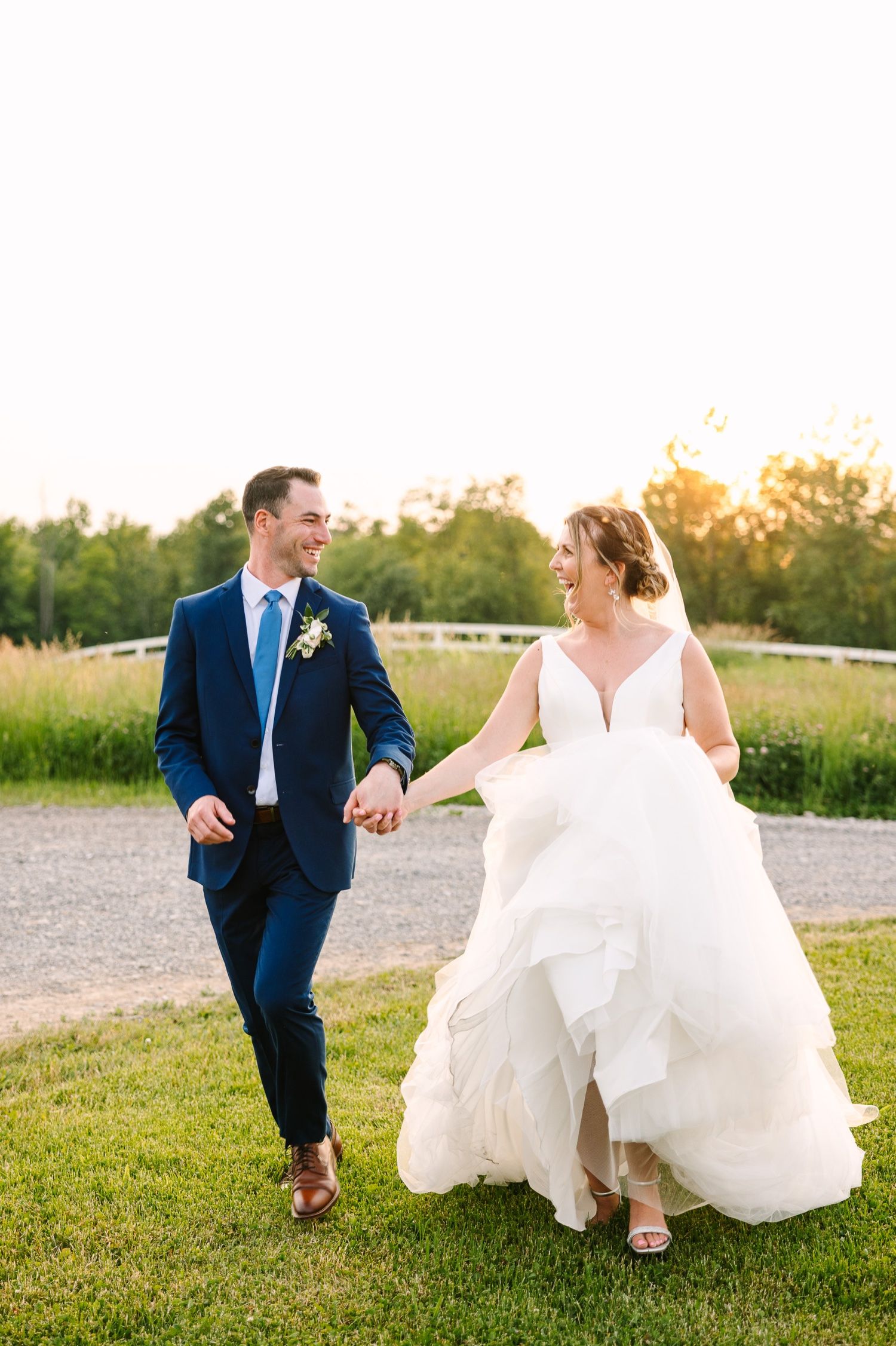 A newlywed couple runs playfully through a grassy field during golden hour.