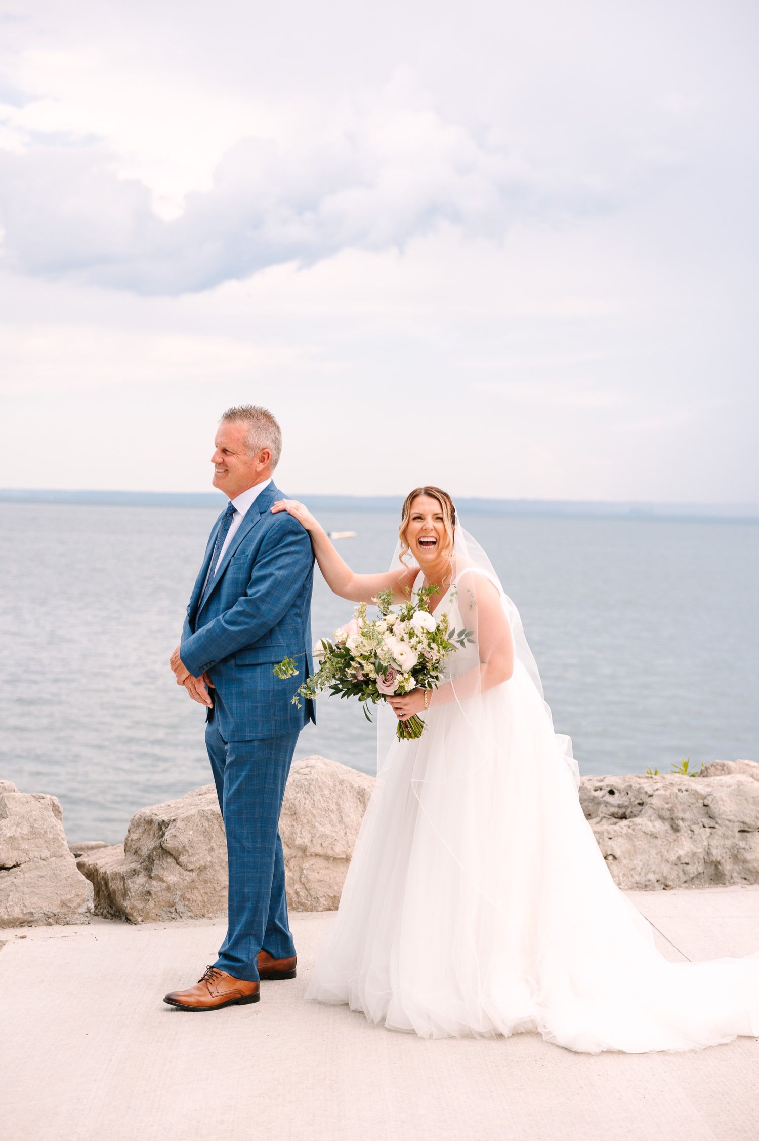 A couple shares a playful first look moment against a lakefront backdrop.