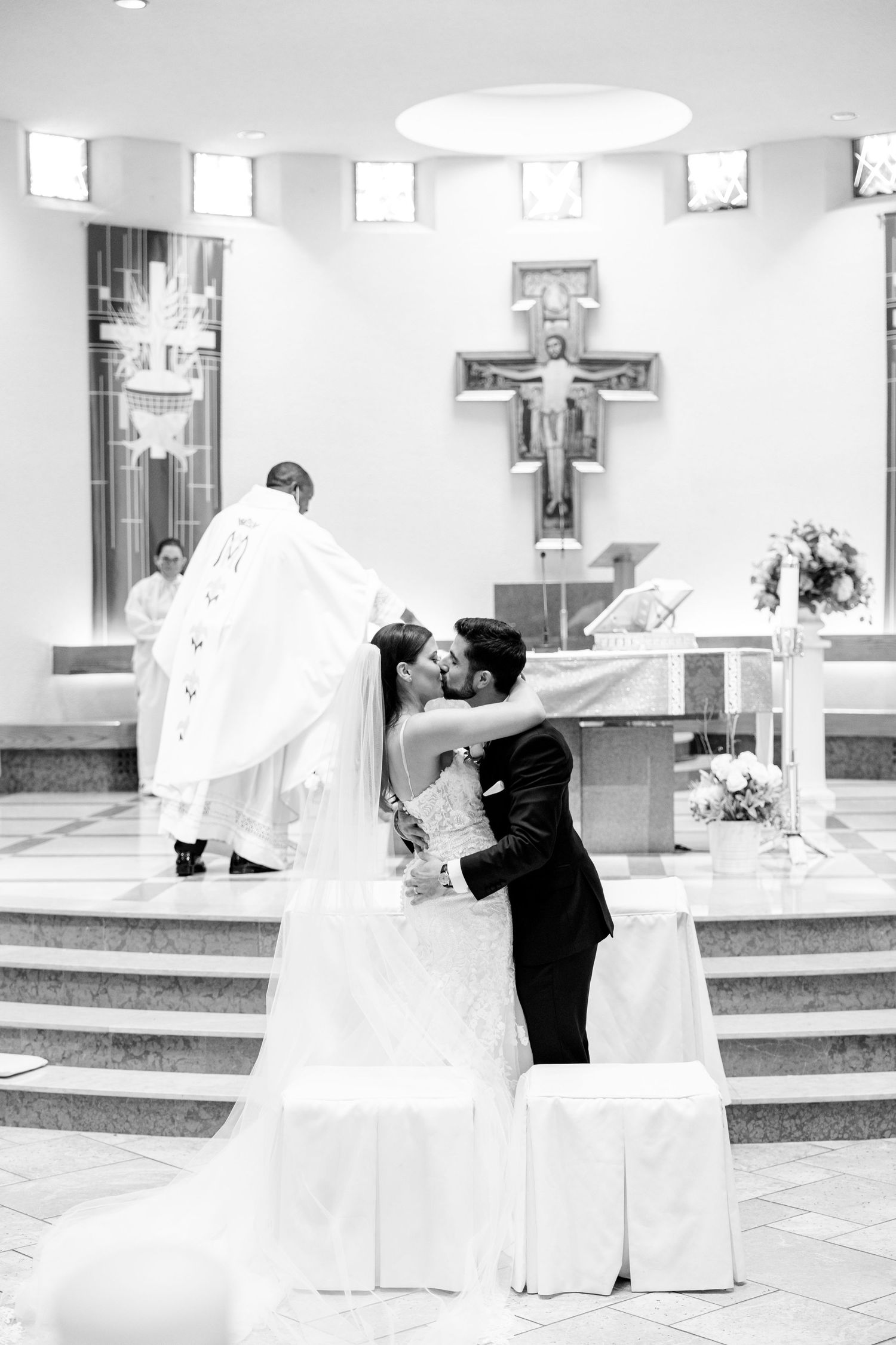 A dramatic black and white photo of newlyweds kissing at the church altar with priest in background.