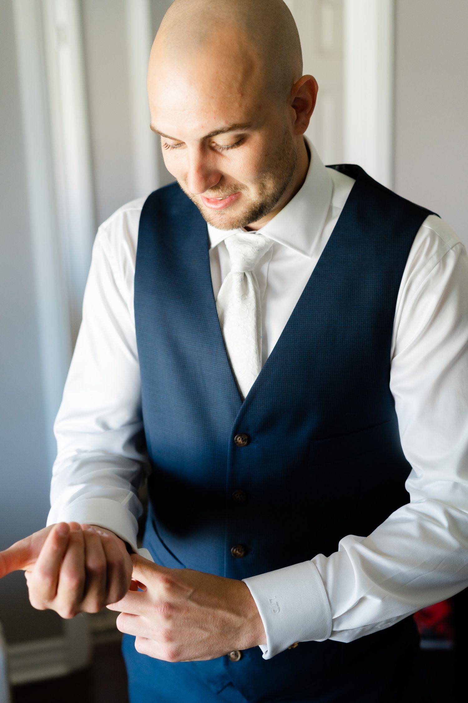 Groom in a navy blue vest adjusts his white collar while getting ready.