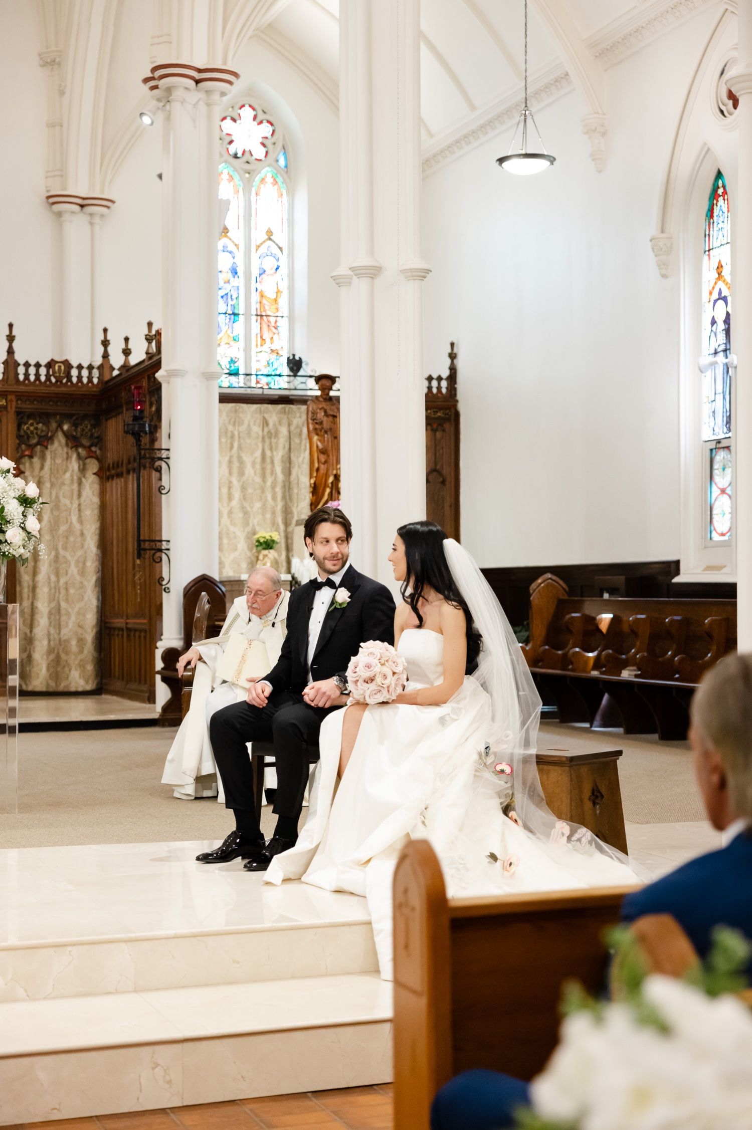 Wedding ceremony in a bright white church with couple seated on altar steps surrounded by stained glass windows.
