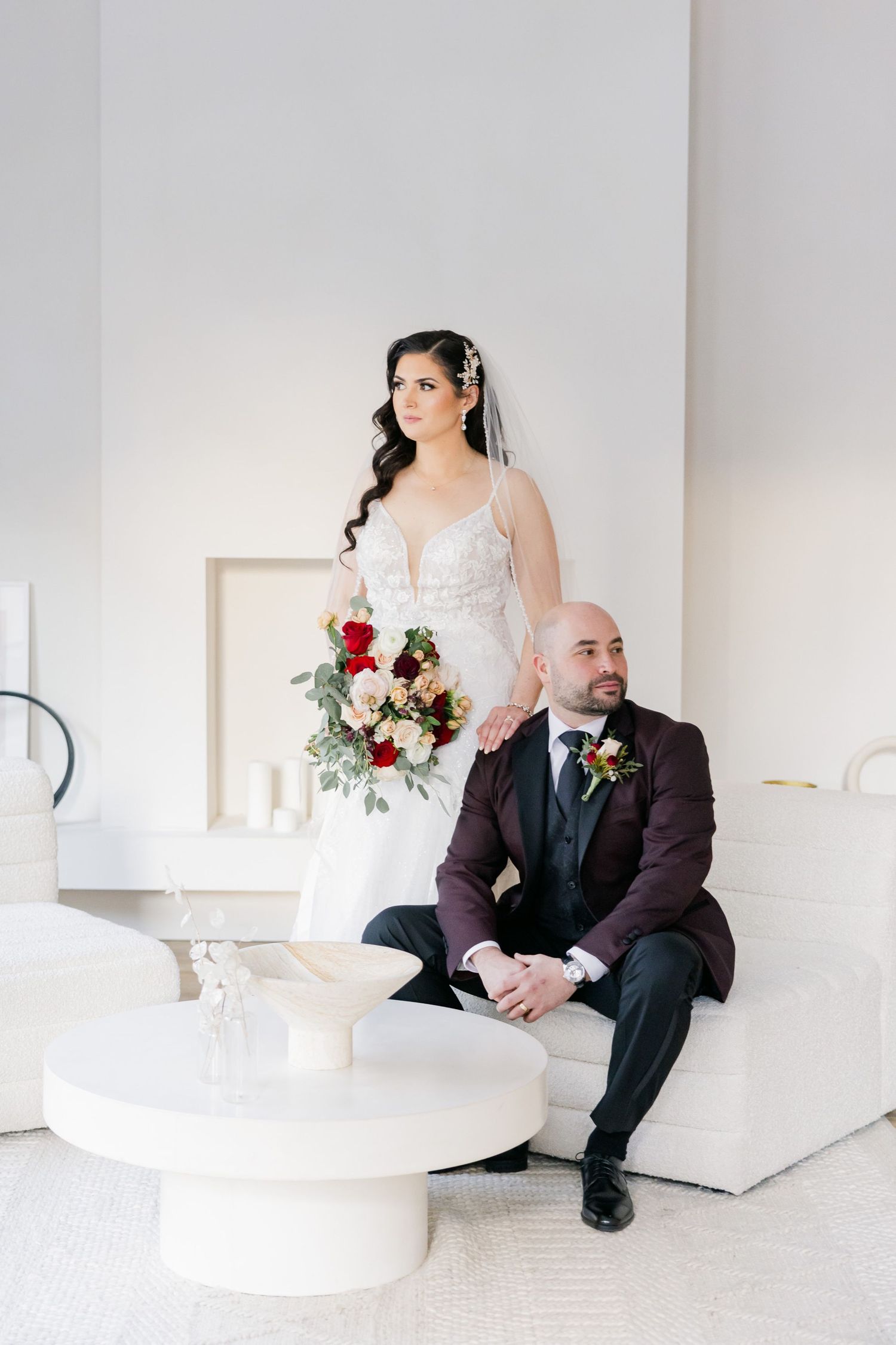 A wedding couple poses in a modern white room with a red and white bouquet.