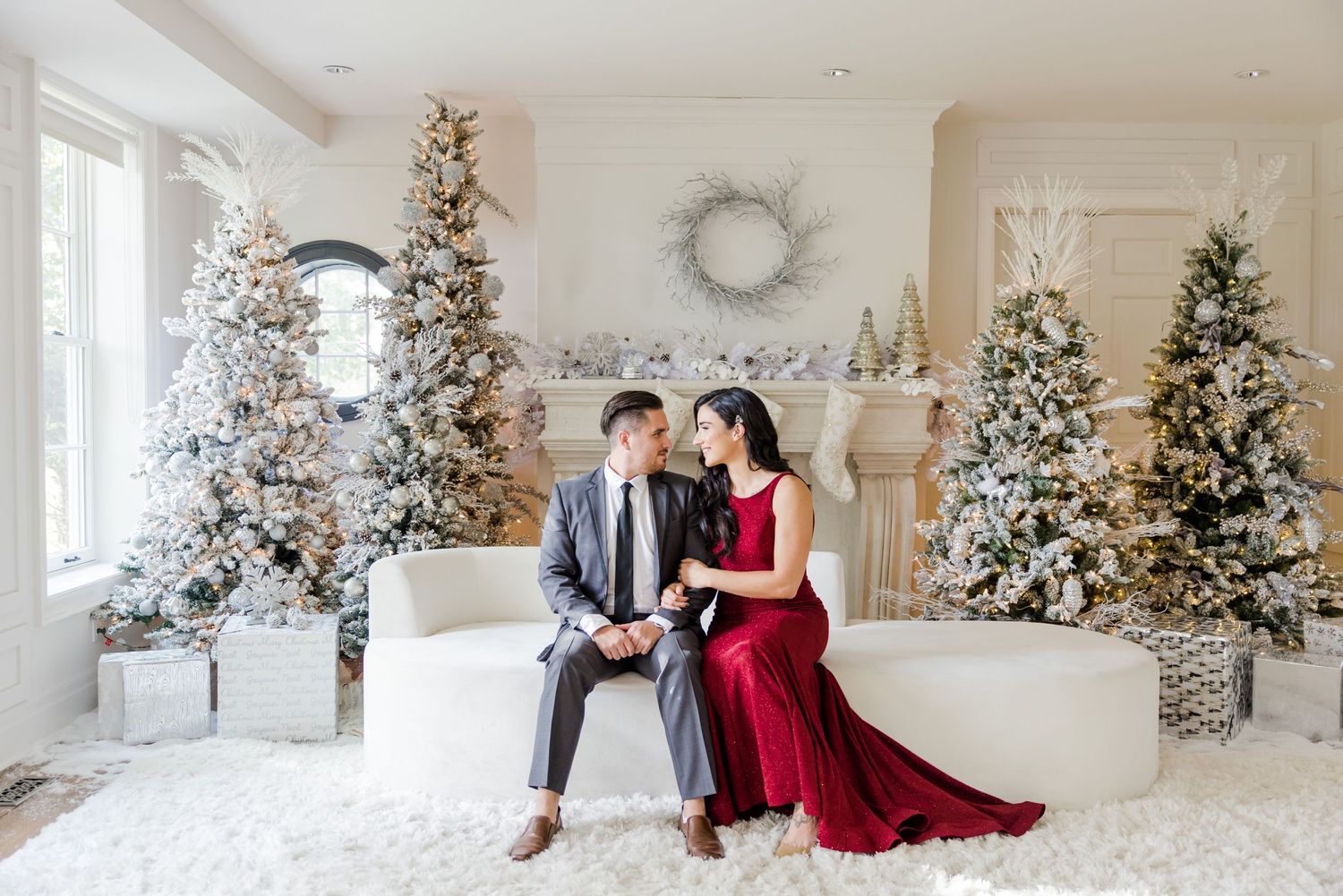 Couple in formal attire seated on white bench surrounded by decorated Christmas trees in bright white room.