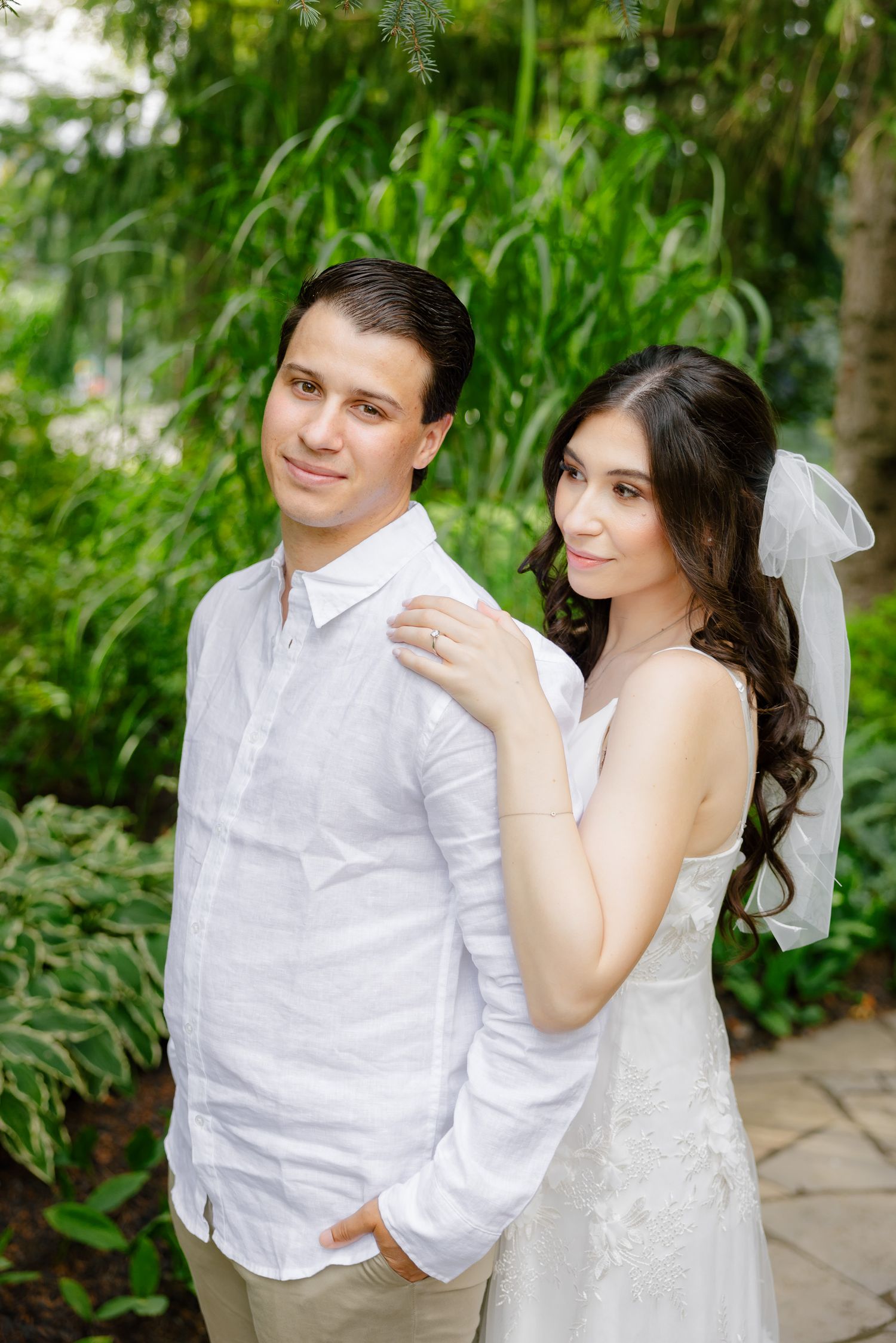 A wedding couple stands in a garden setting wearing coordinated white outfits.