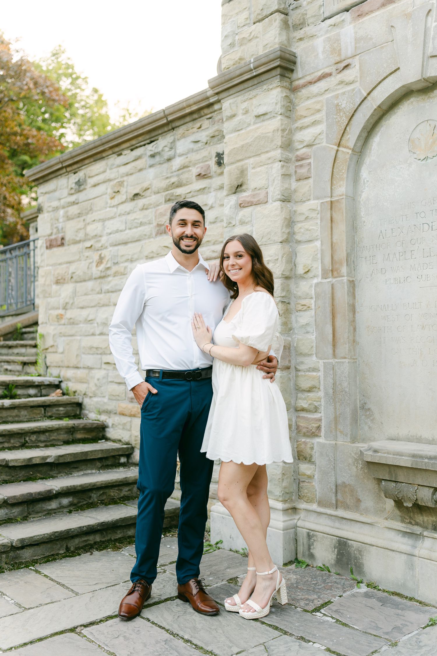 Elegant couple poses against stone wall architecture in formal garden setting.