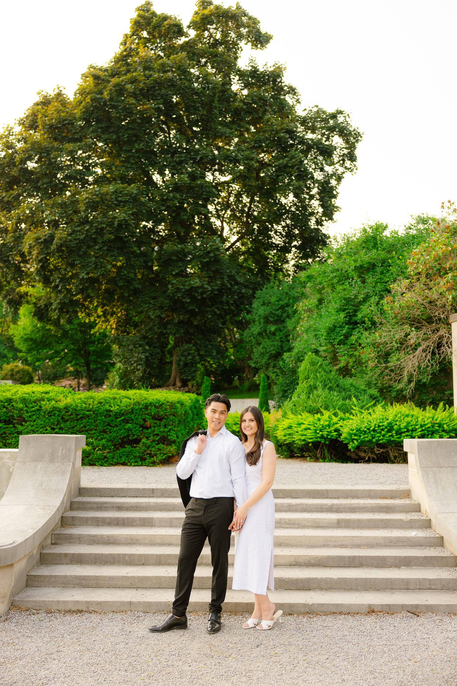 Couple poses for a portrait on curved stone steps in a manicured garden with towering trees.