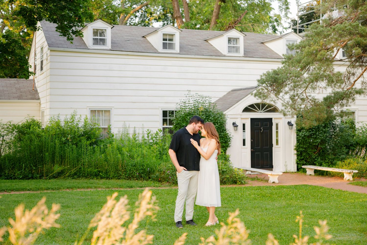 Intimate moment between couple outside classic white house with dormer windows and landscaped garden.