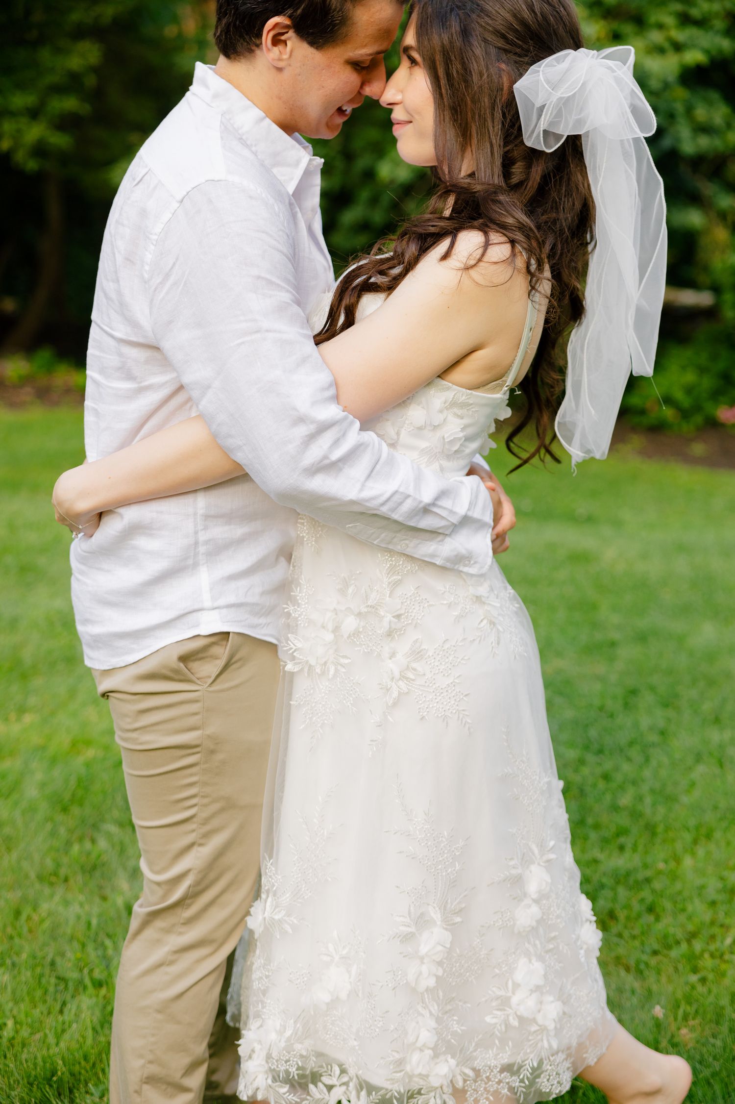 Intimate moment between couple in wedding attire standing on grass in a garden setting.