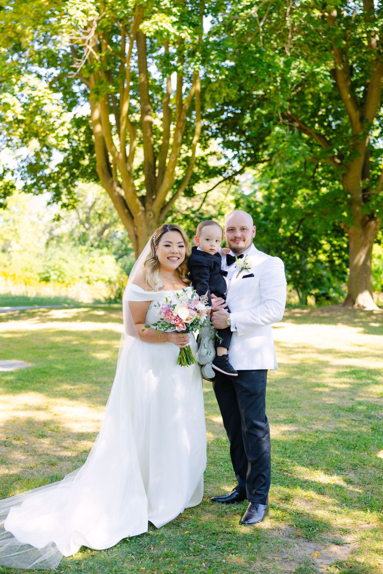 Elegant outdoor wedding portrait with couple and toddler under sunlit trees.