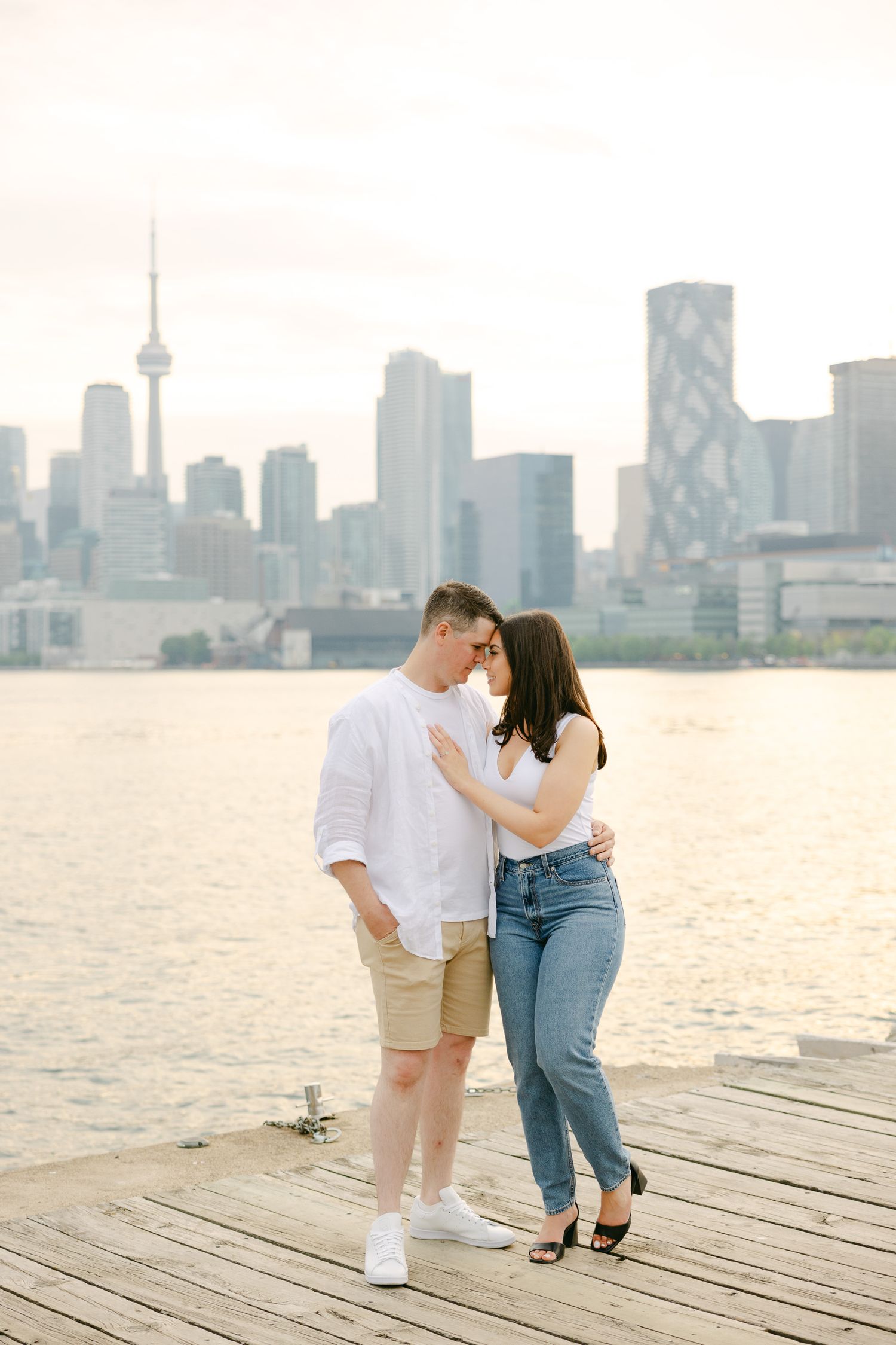 Couple in casual summer outfits sharing a romantic moment on waterfront deck with Toronto skyline behind.