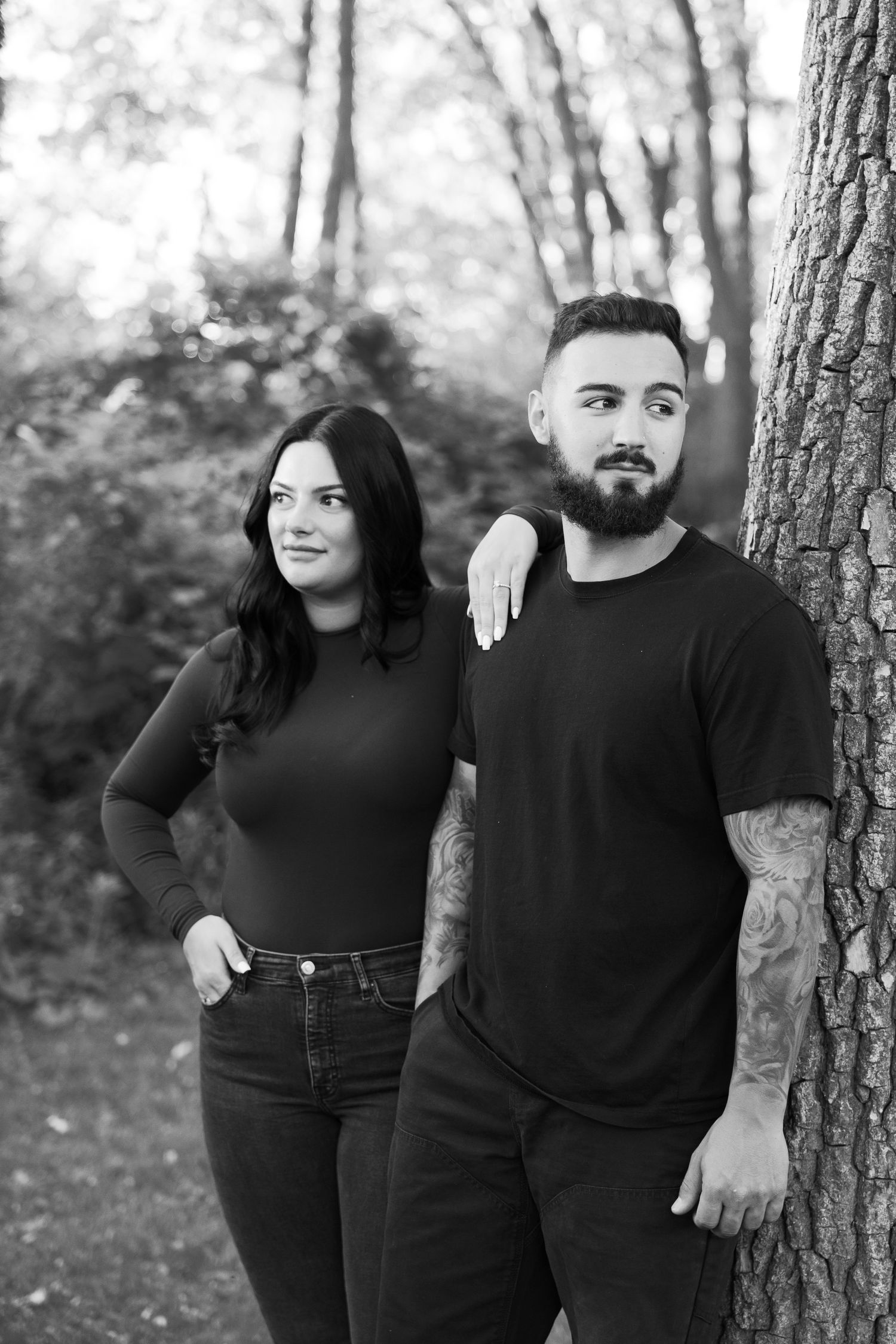 Couple in black outfits posing together against a tree trunk in black and white outdoor portrait.