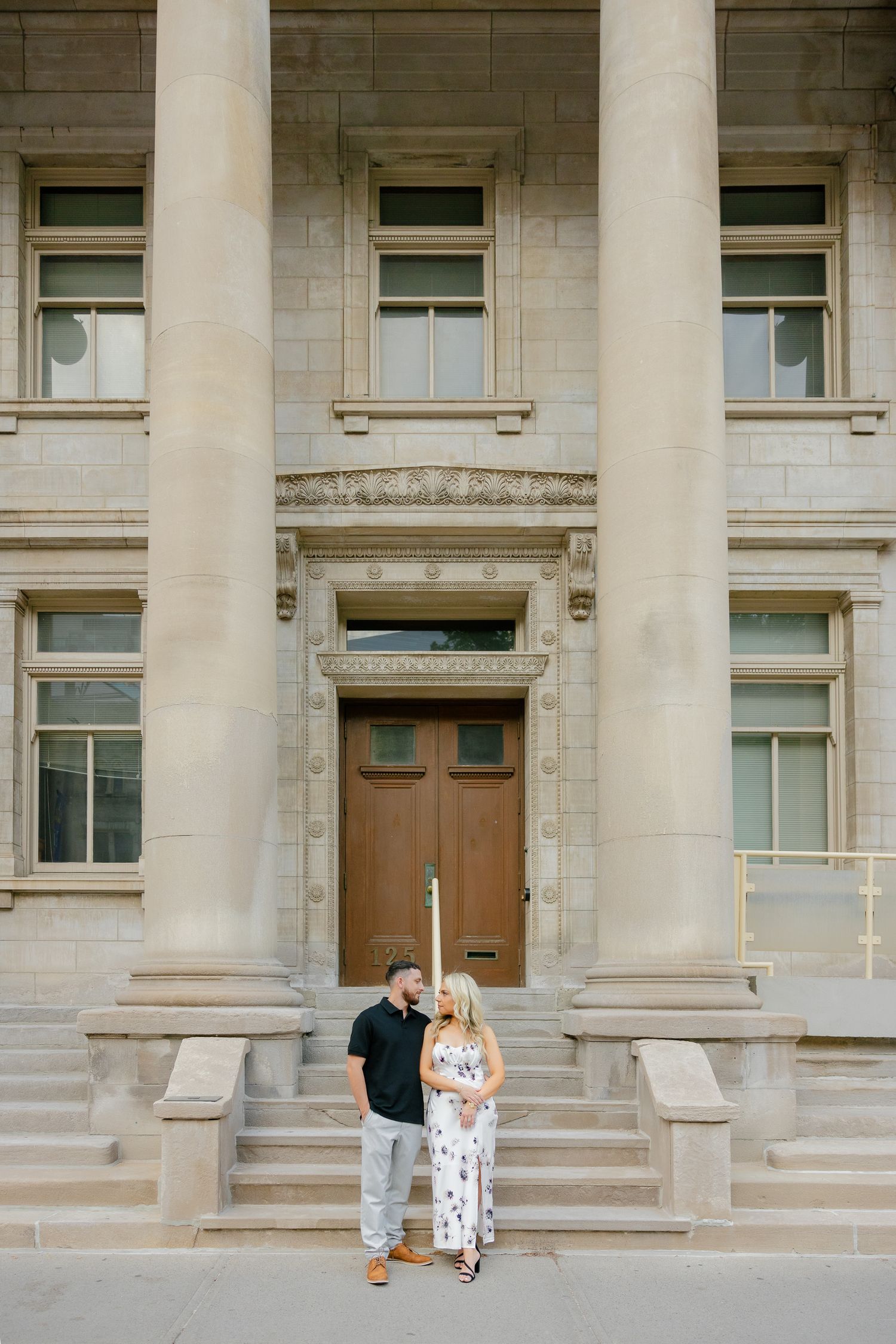 A couple stands together on ornate stone steps of a neoclassical building entrance.