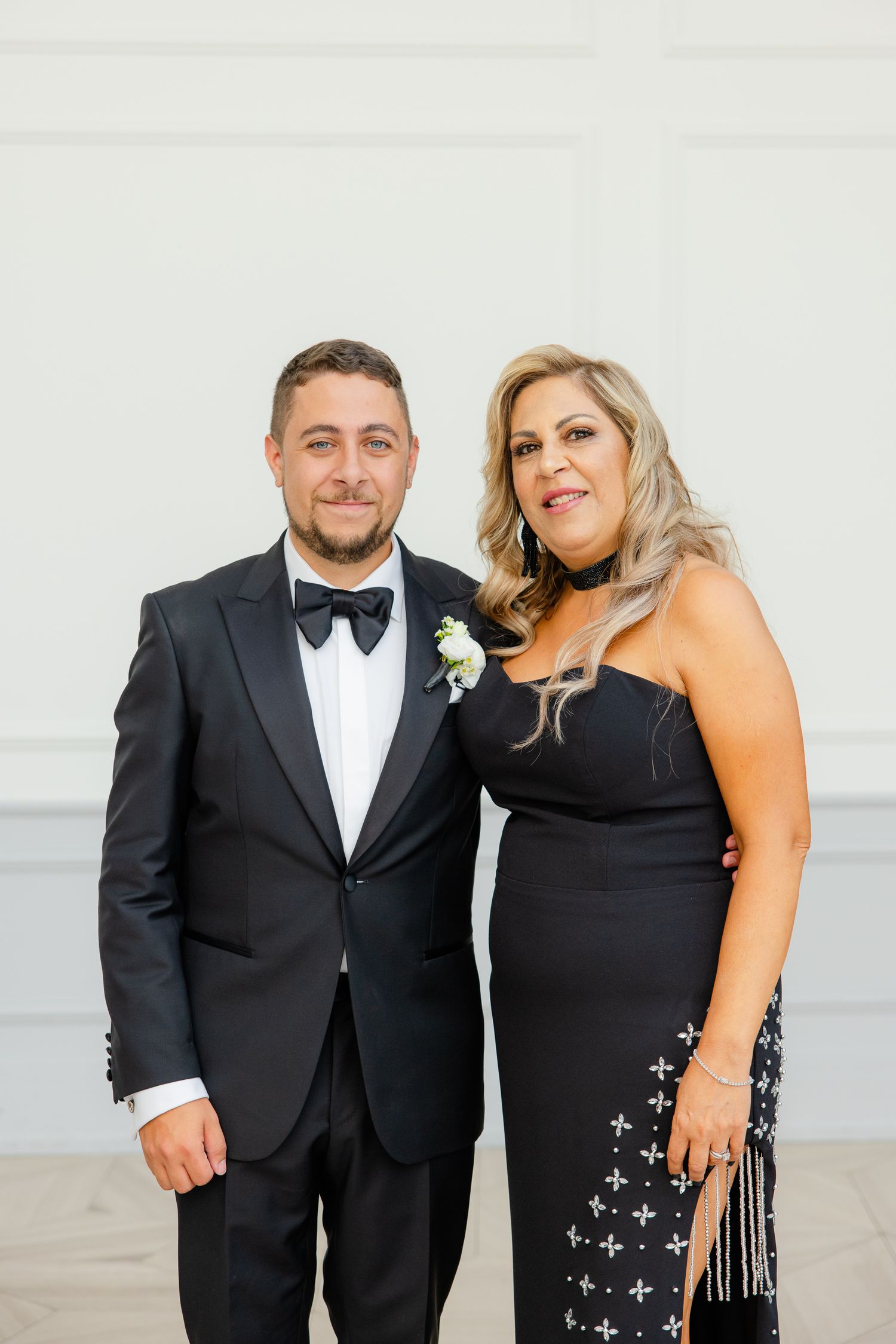 A couple in formal black attire pose together at a wedding celebration against a light background.